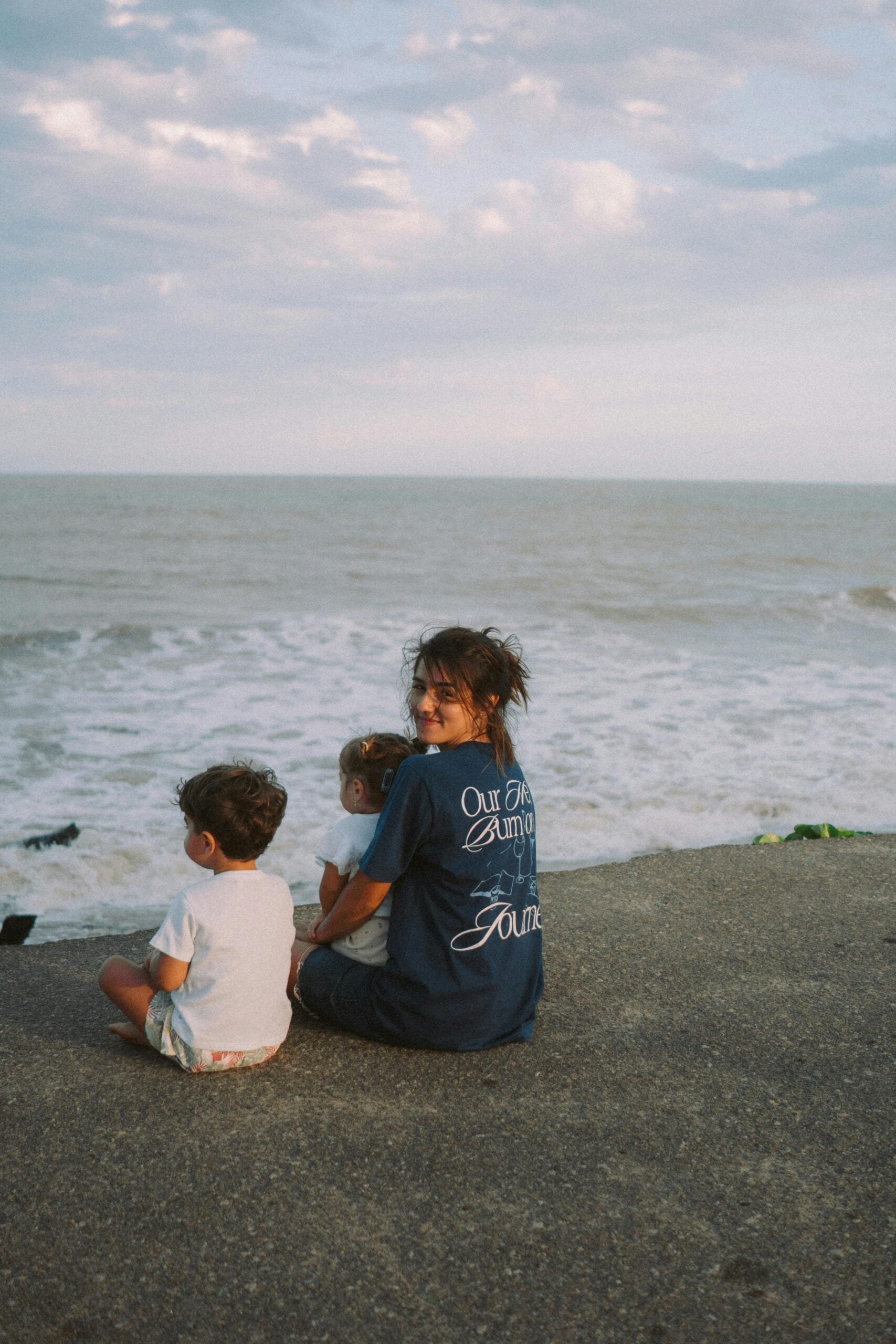 A woman and two young children sit on a concrete ledge by the ocean, facing the water. The woman smiles at the camera, while the children look toward the waves under a partly cloudy sky.