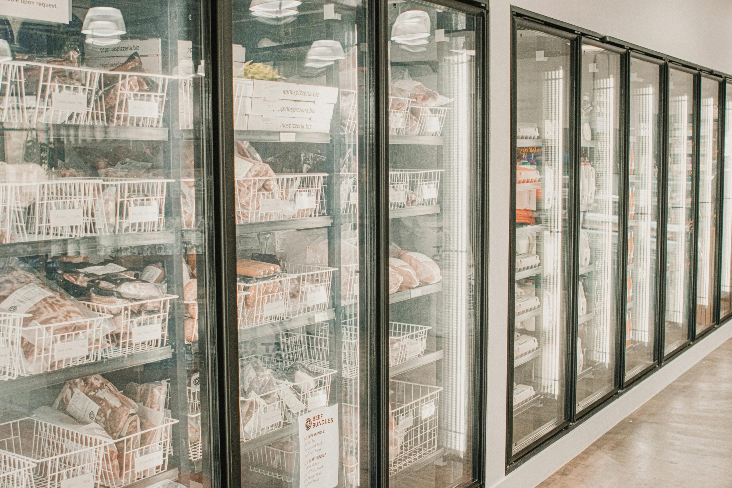 Glass-door freezers in a grocery store, filled with baskets containing packaged frozen foods. The freezers are well-lit and line the wall, with the polished floor reflecting the lights above.