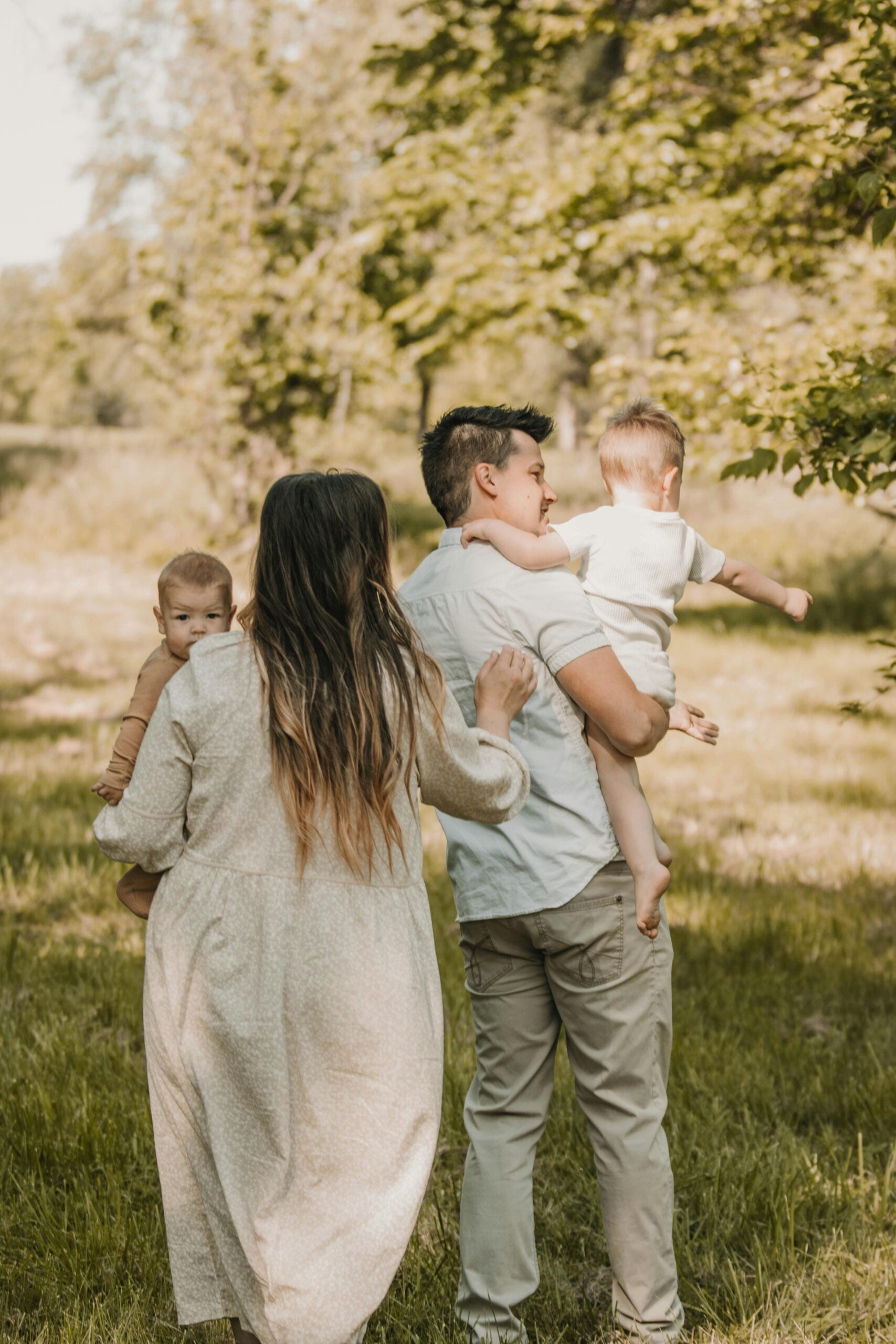 A family of four walks outdoors on a grassy path. The mother holds a baby, and the father carries a toddler. They are walking away from the camera surrounded by trees and sunlight.