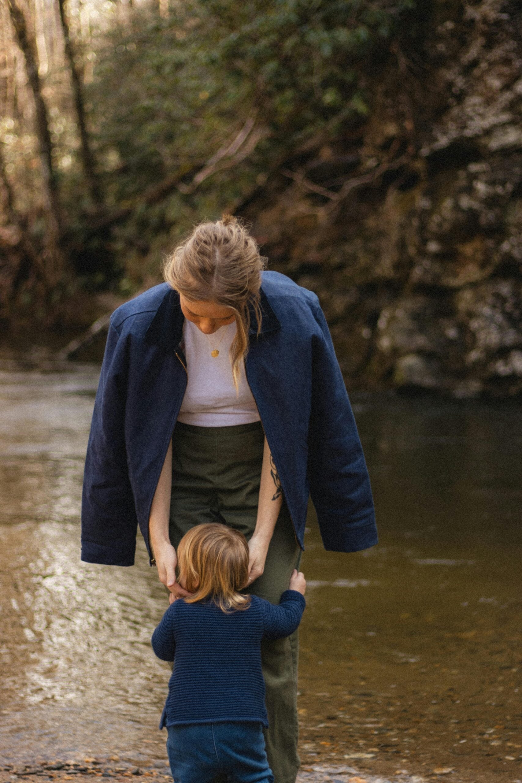 A woman stands by a creek, looking down as a small child hugs her legs. Both wear blue clothing and are surrounded by trees and nature.
