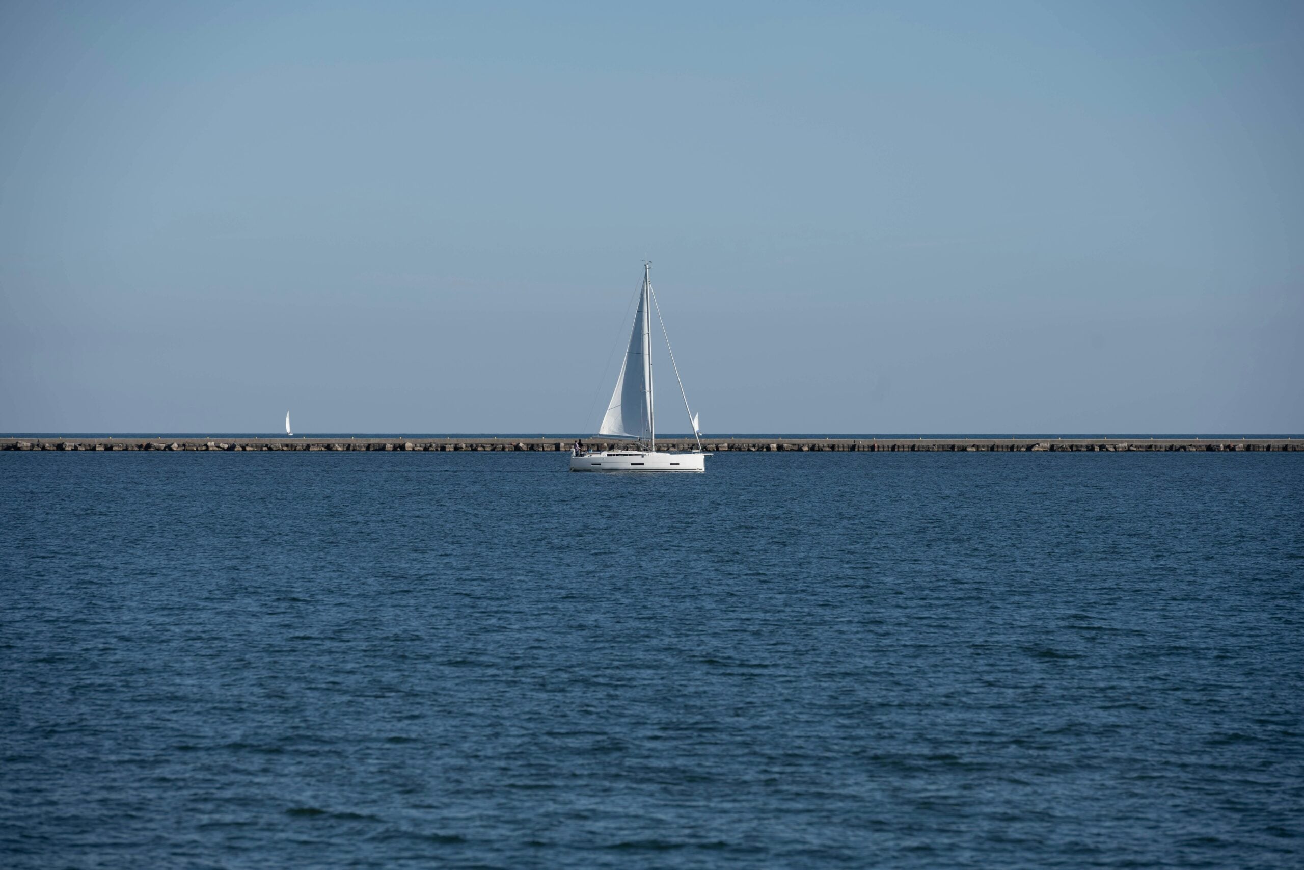 A white sailboat sails on calm blue water under a clear sky, with a long stone breakwater in the background and a smaller sailboat visible in the distance.