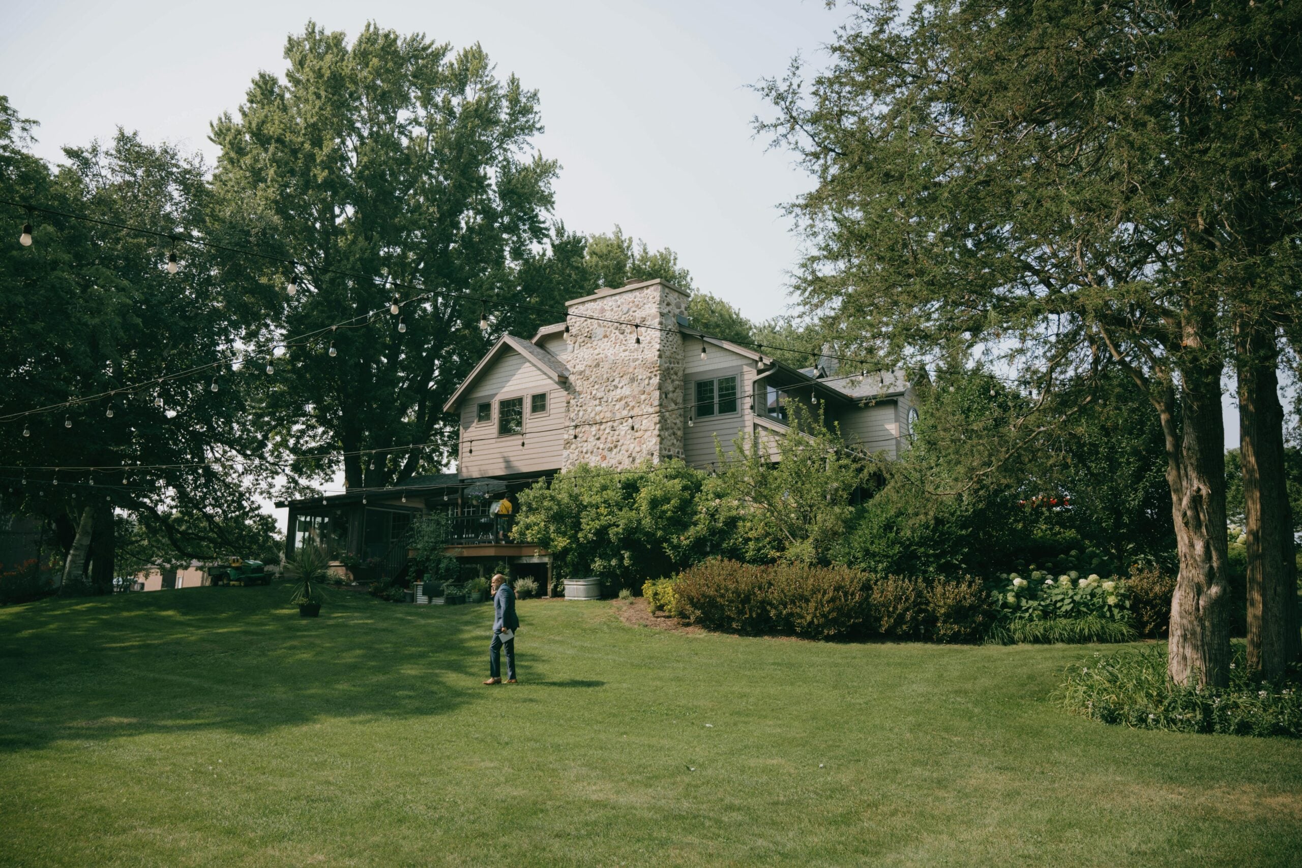 A large, gray stone house with a chimney sits surrounded by lush trees and greenery. A person stands on the expansive, well-manicured lawn under string lights on a bright, clear day.