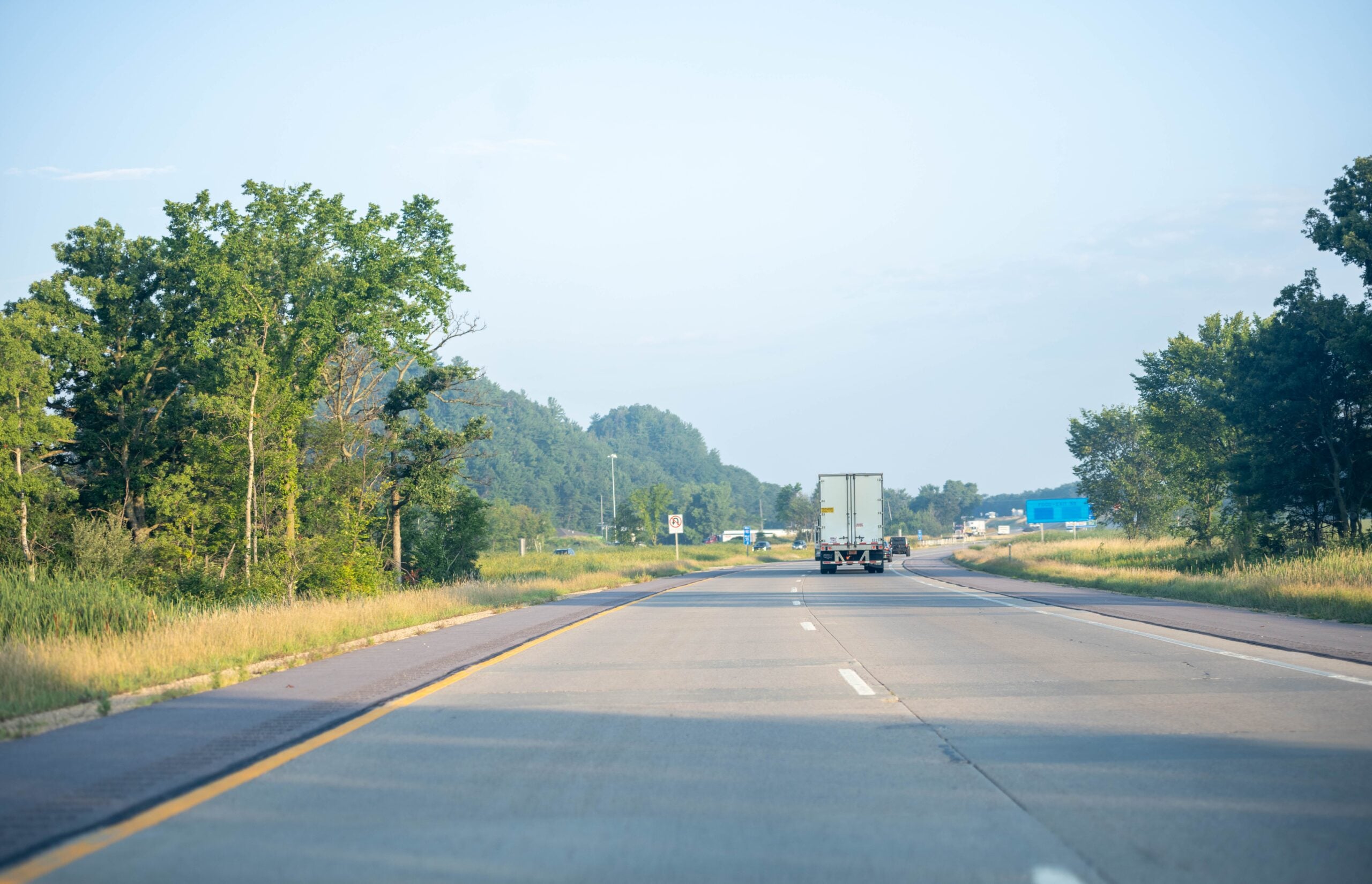 A truck drives down an empty highway surrounded by green trees and grass under a clear blue sky on a sunny day.