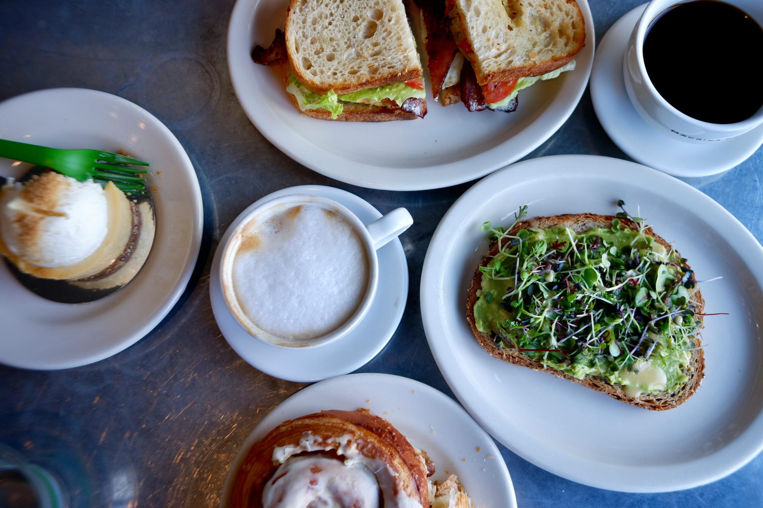 A table with a latte, black coffee, sandwiches, avocado toast with microgreens, a dessert with ice cream, and a cinnamon roll on white plates.