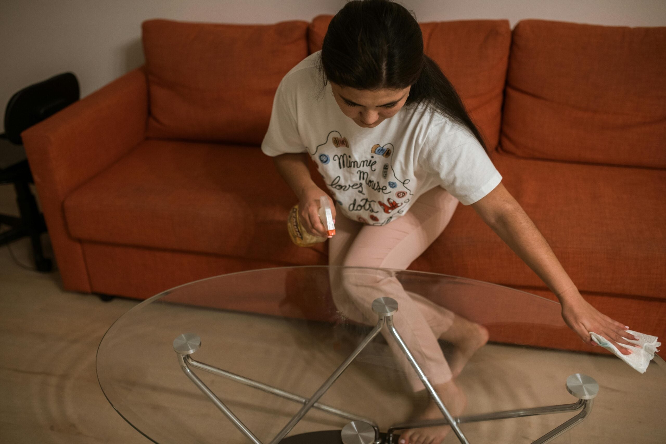 A woman is cleaning a glass coffee table with a spray bottle and cloth in a living room with an orange sofa in the background.