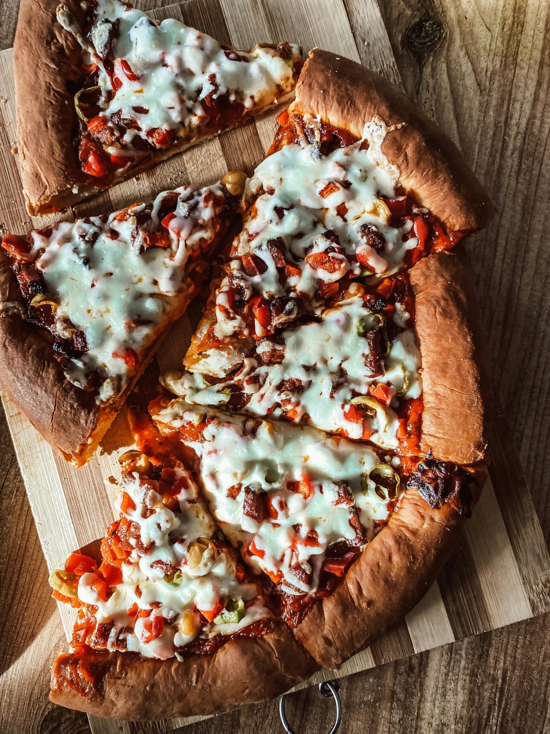 A close-up of a sliced pizza with melted cheese, peppers, and other toppings on a wooden cutting board atop a rustic wooden table.