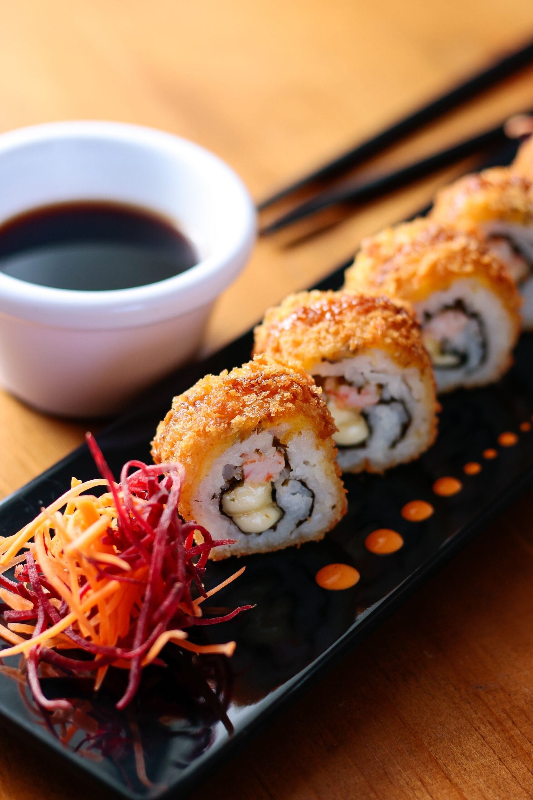 A close-up of crispy sushi rolls topped with sauce, served on a black plate with a colorful shredded vegetable garnish and a small bowl of soy sauce in the background.