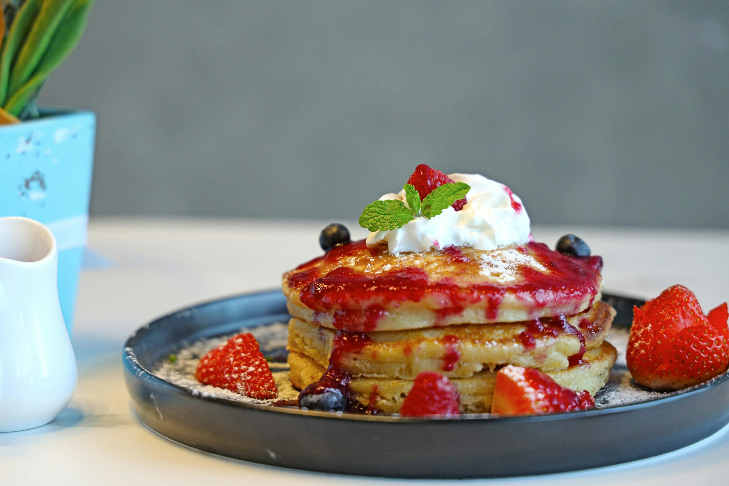 A stack of pancakes topped with whipped cream, berry sauce, a mint leaf, and surrounded by strawberries and blueberries on a dark plate. A small pitcher and a blue pot are in the background.