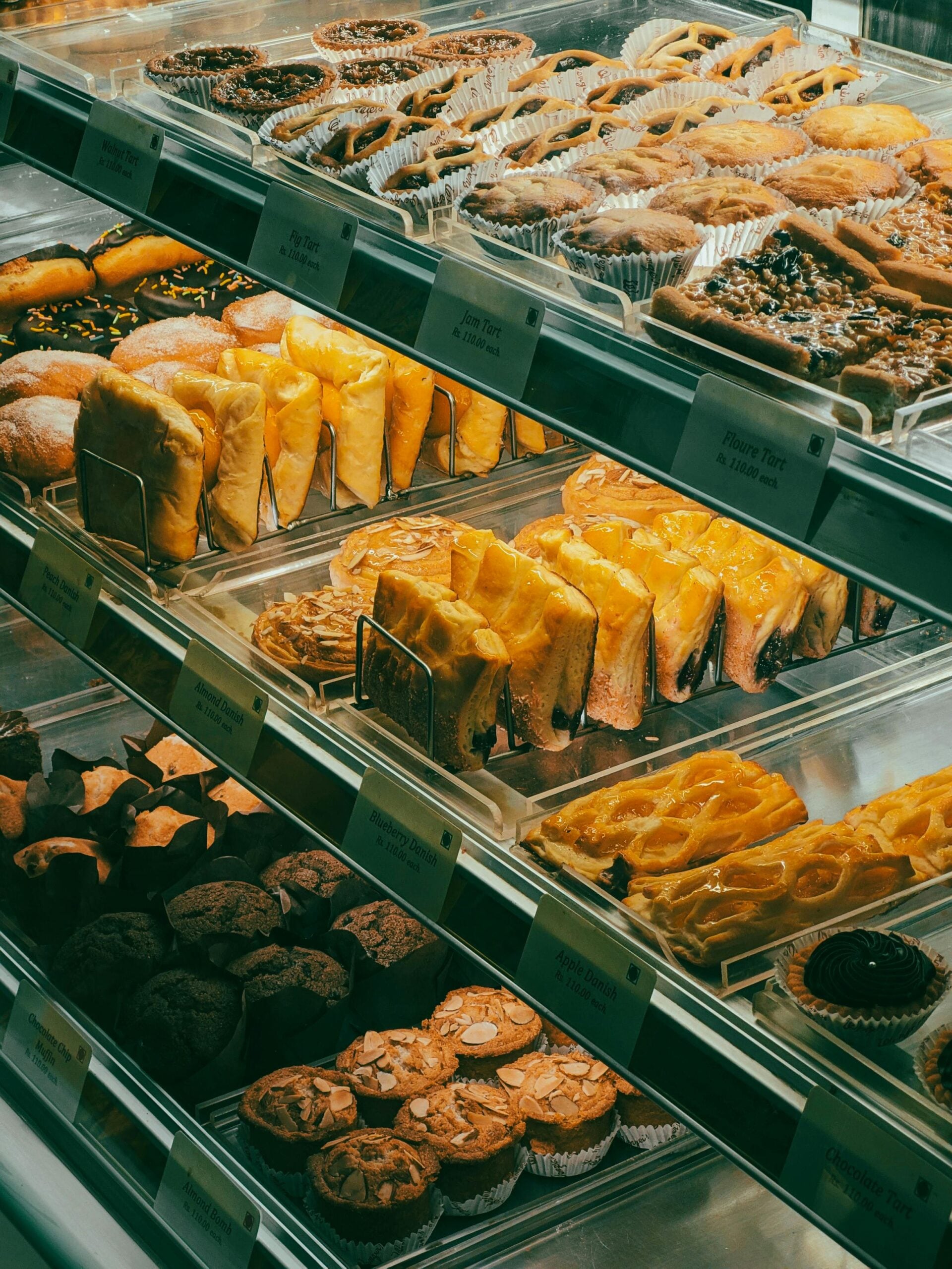 A bakery display case filled with a variety of pastries, tarts, and cakes neatly arranged on metal shelves, with labels in front of each dessert.