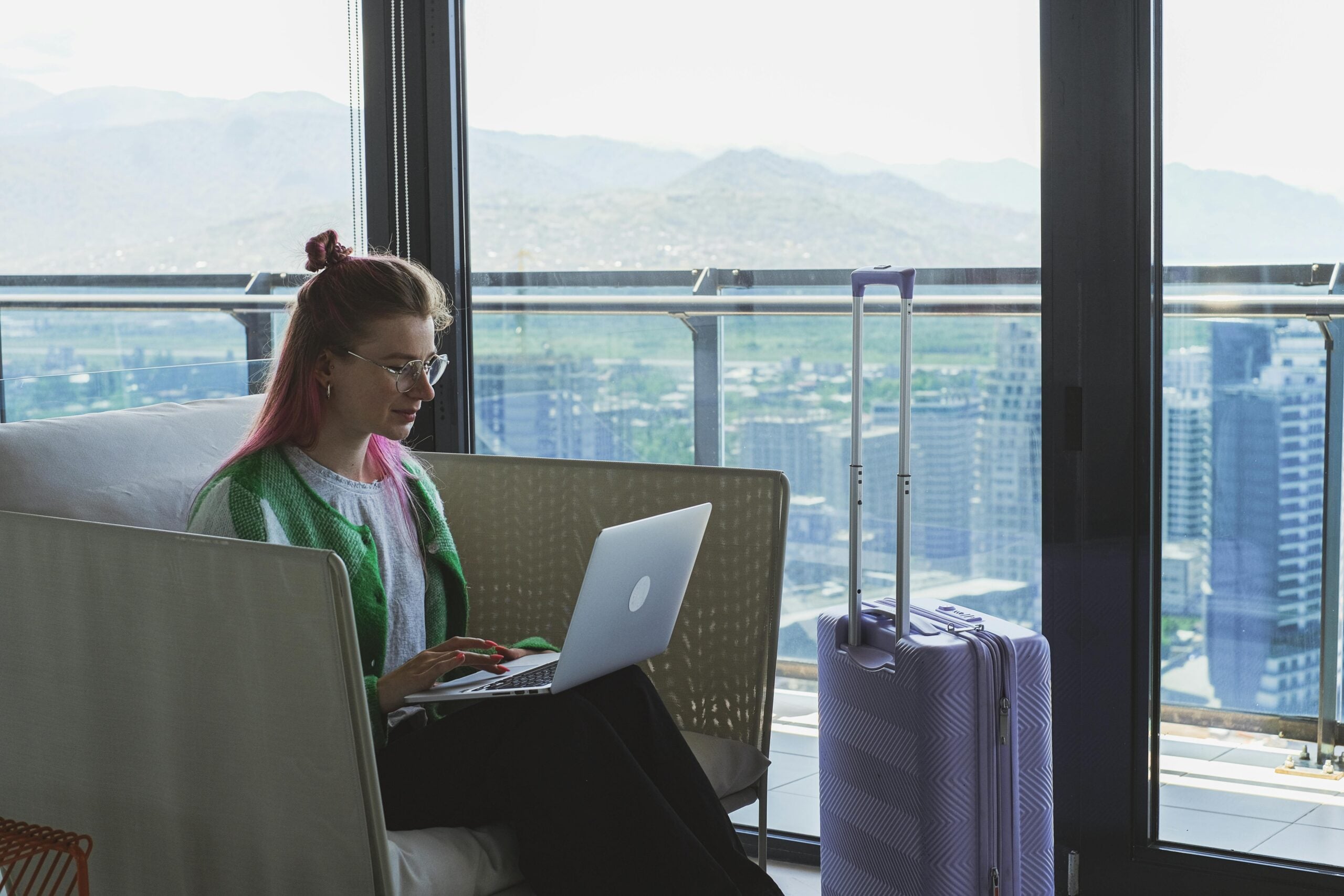 A woman with pink hair sits on a couch using a laptop next to a purple suitcase in a modern room with large windows overlooking a cityscape and mountains.