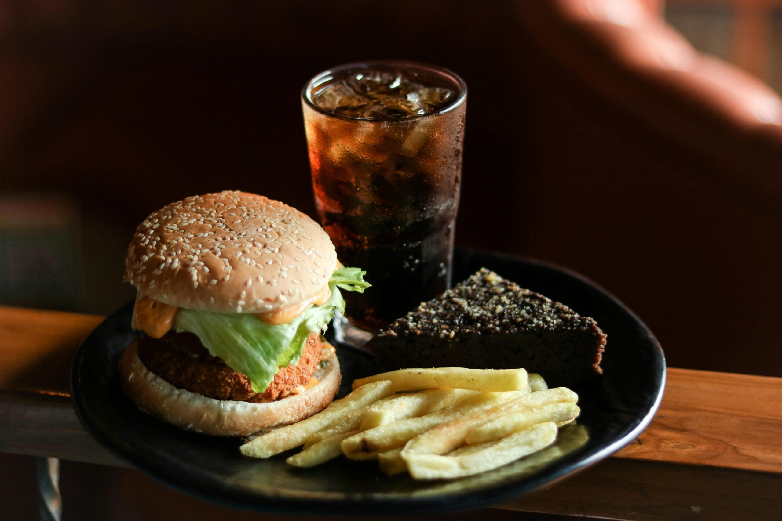 A black plate with a burger containing lettuce, a serving of French fries, a slice of chocolate cake with sprinkles, and a glass of cola filled with ice.
