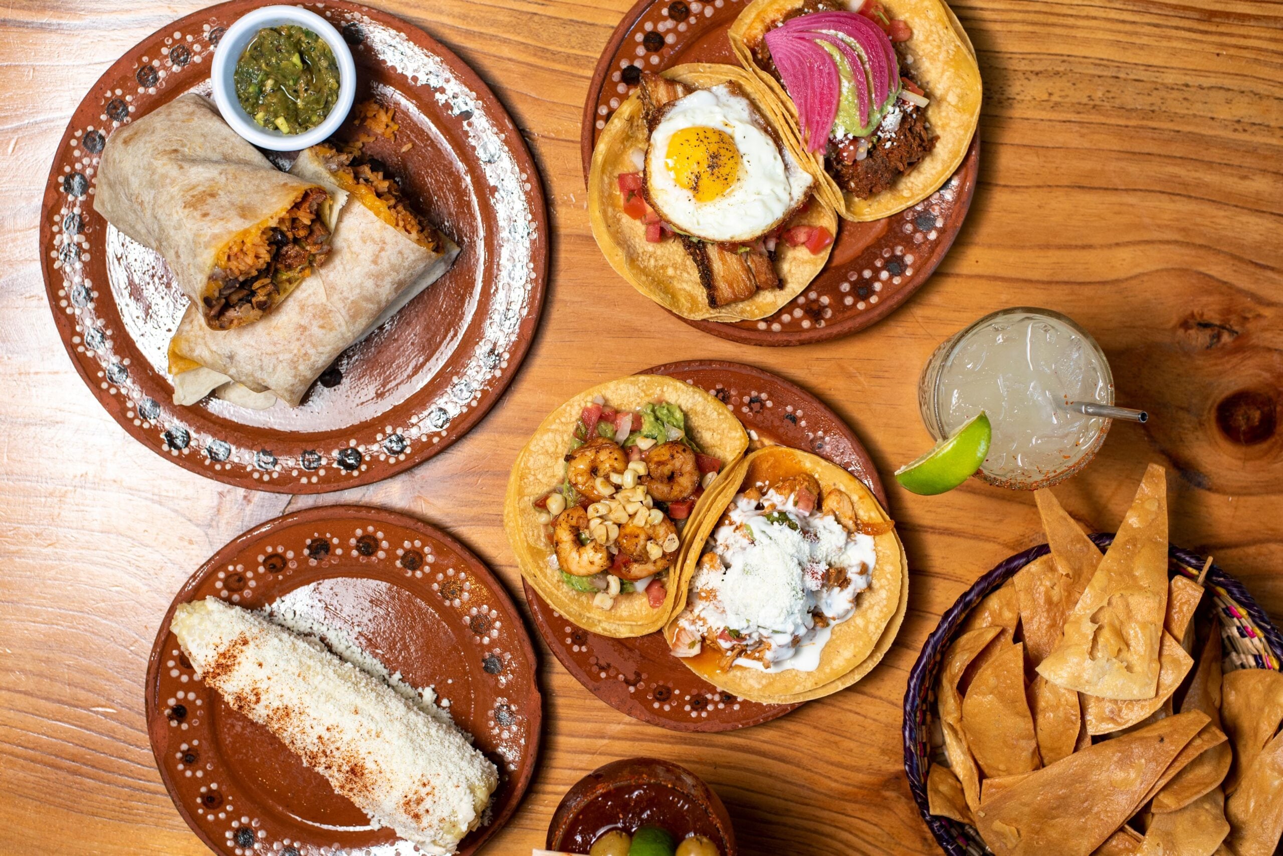 Overhead view of Mexican dishes on a wooden table, including a burrito, three tacos with various toppings, elote (grilled corn), tortilla chips with salsa, and a cocktail with a lime wedge.