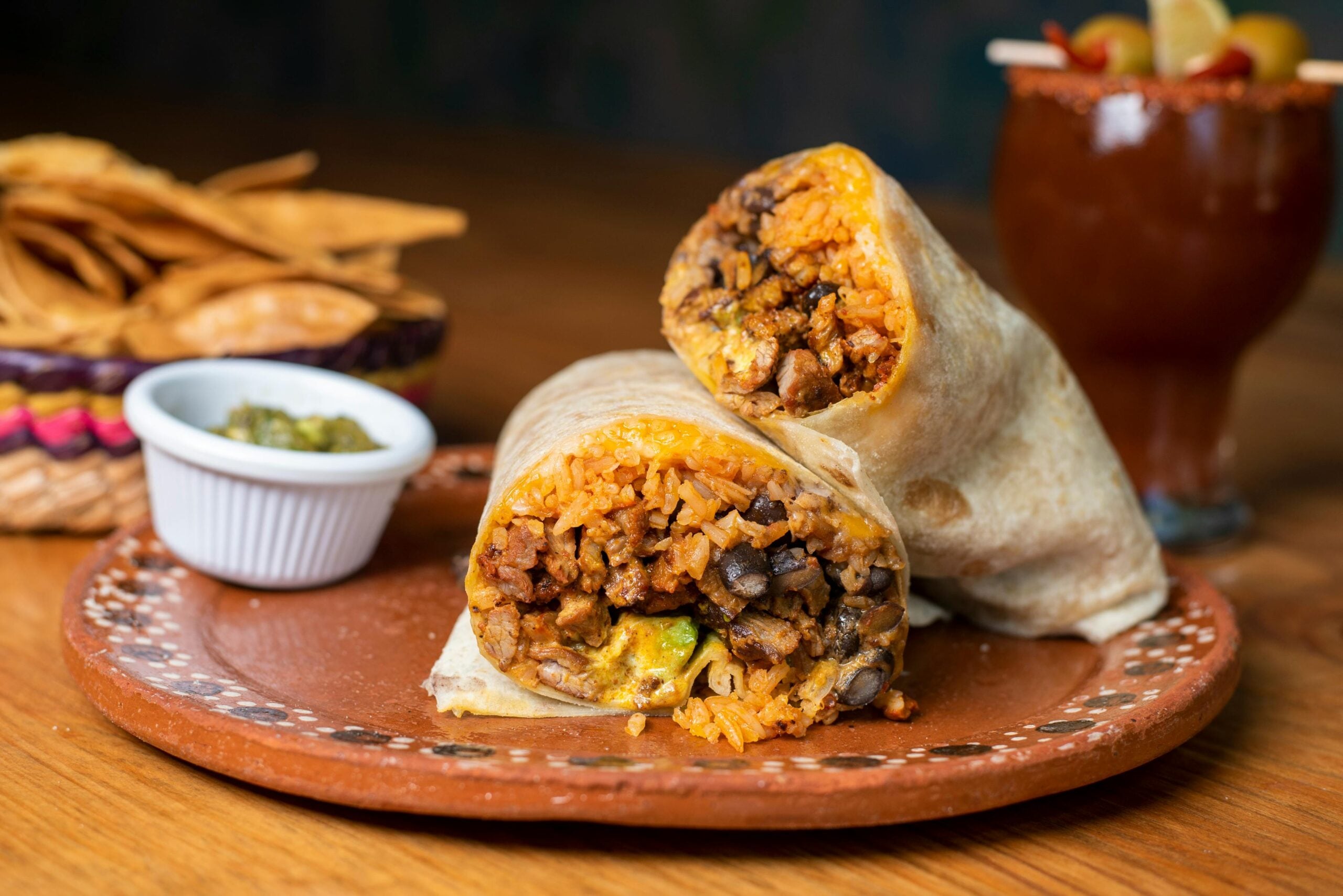 A burrito cut in half shows its filling of rice, beans, meat, and vegetables on a clay plate. Nearby are tortilla chips in a basket, a small dish of salsa, and a dark drink with fruit garnish in the background.