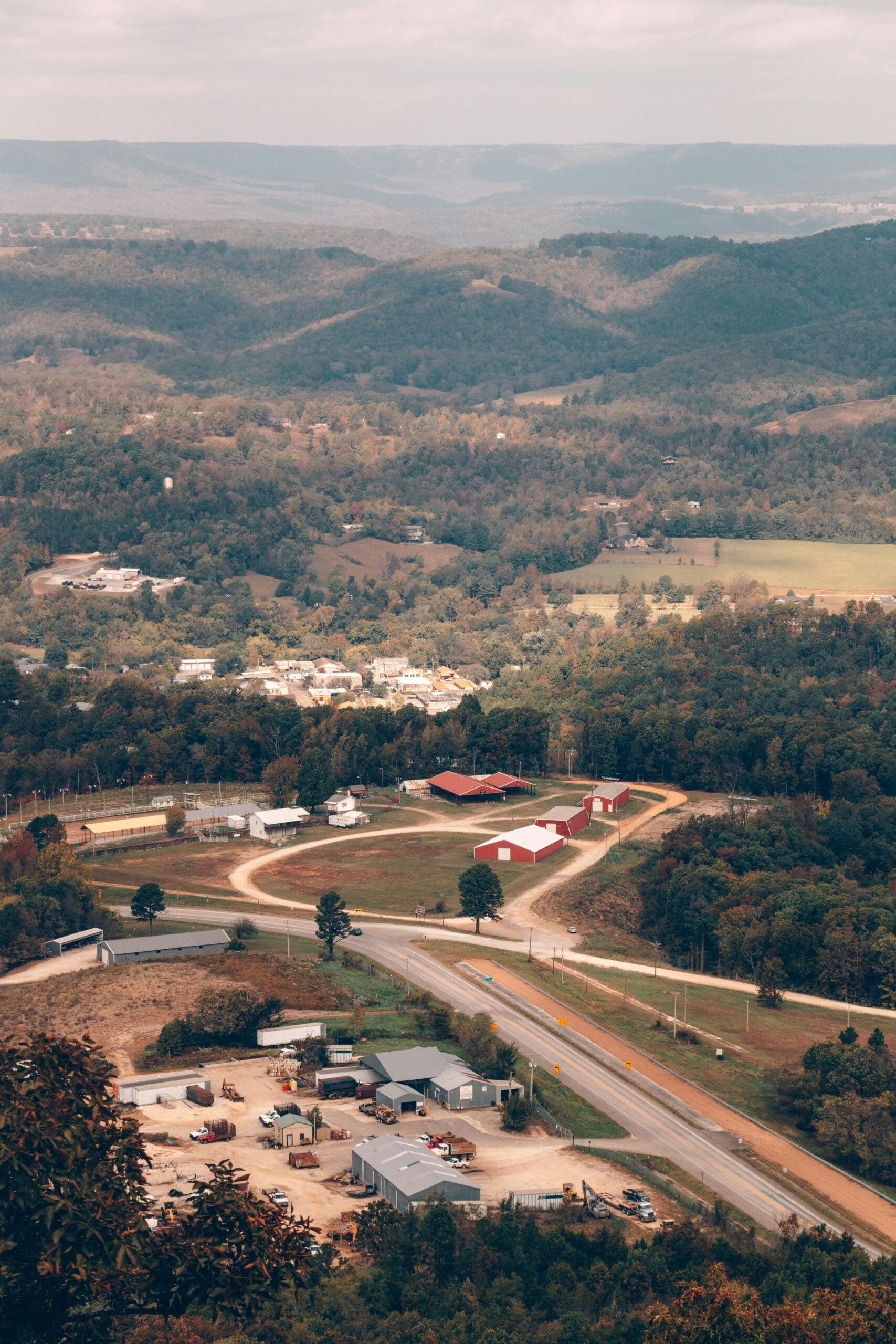 An aerial view of a rural area with small buildings, red-roofed barns, winding roads, and lush green hills in the background under a cloudy sky.