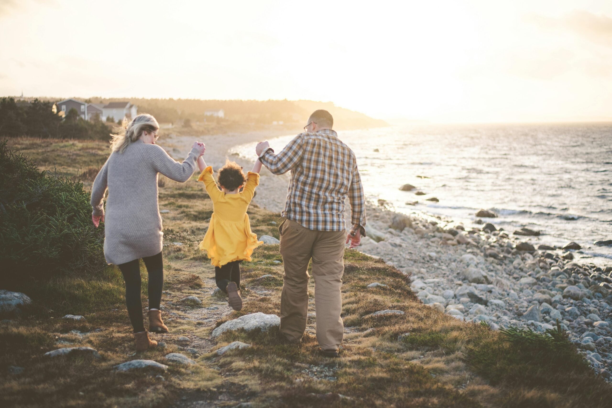 A woman, a man, and a child in a yellow coat walk along a rocky shoreline at sunset. The adults hold the child’s hands, swinging them playfully as they walk away from the camera.