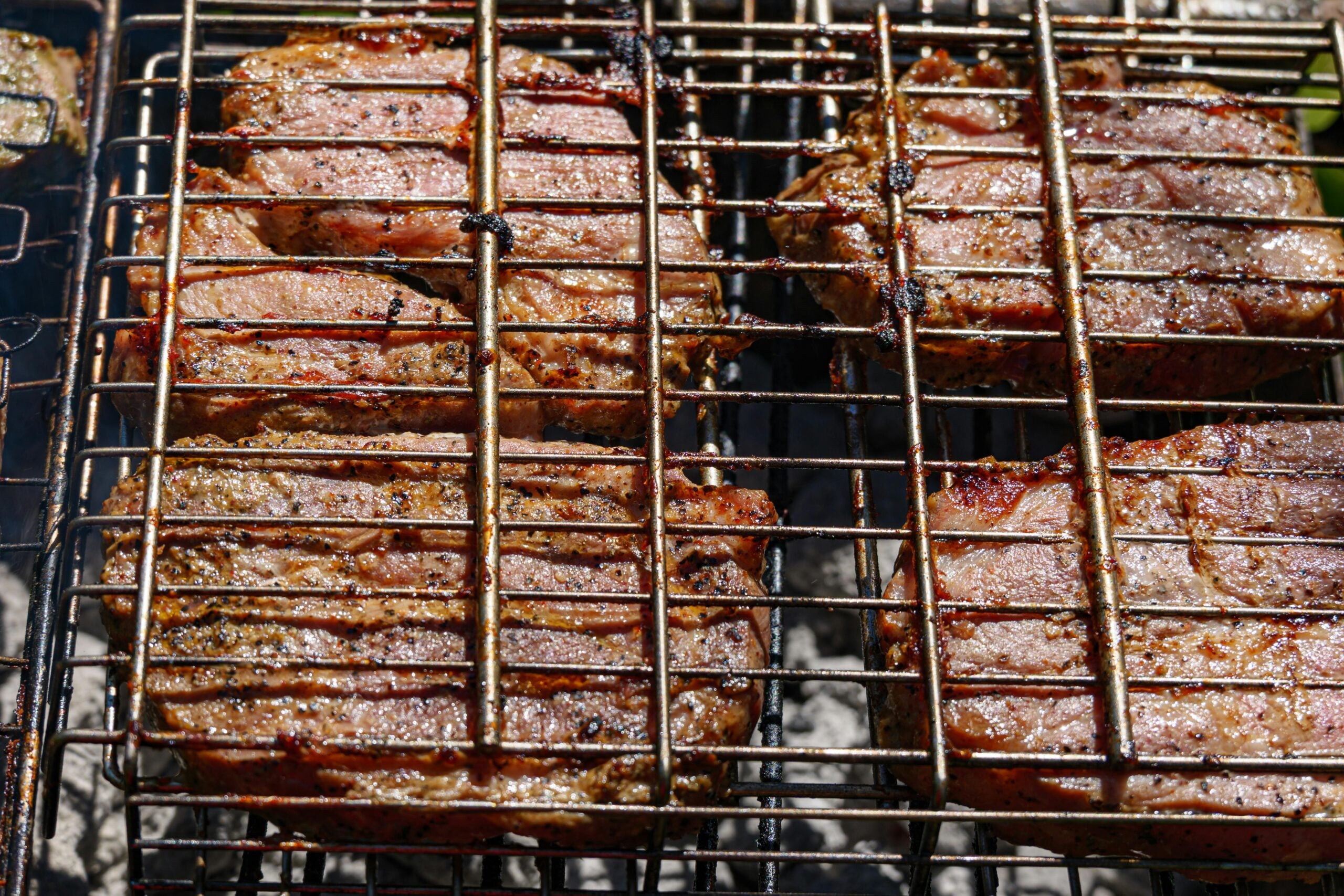 Four seasoned steaks are being grilled on a metal barbecue grill over hot coals, with visible grill marks and a slightly charred, juicy appearance.