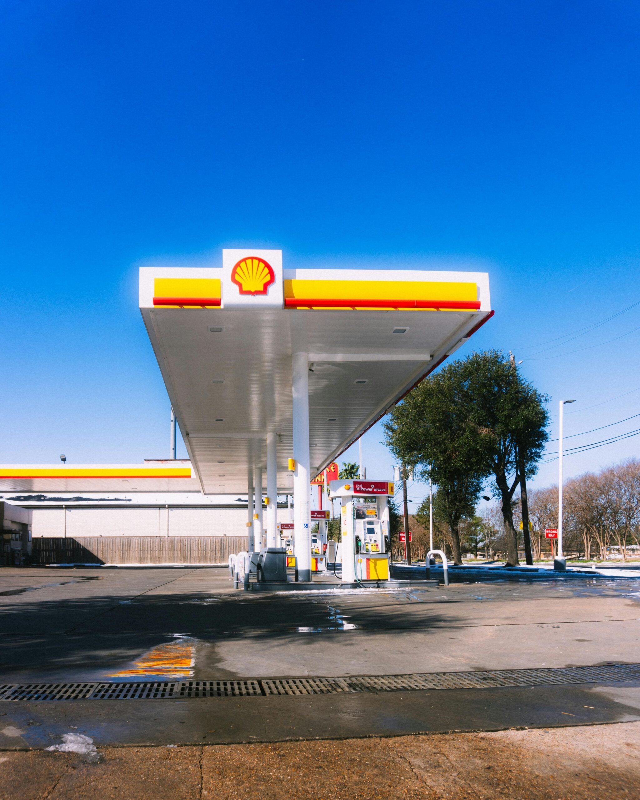 A Shell gas station with a large canopy and fuel pumps sits under a clear blue sky, surrounded by trees and some patches of snow on the ground. The area appears quiet and empty of vehicles and the best day to buy gas.