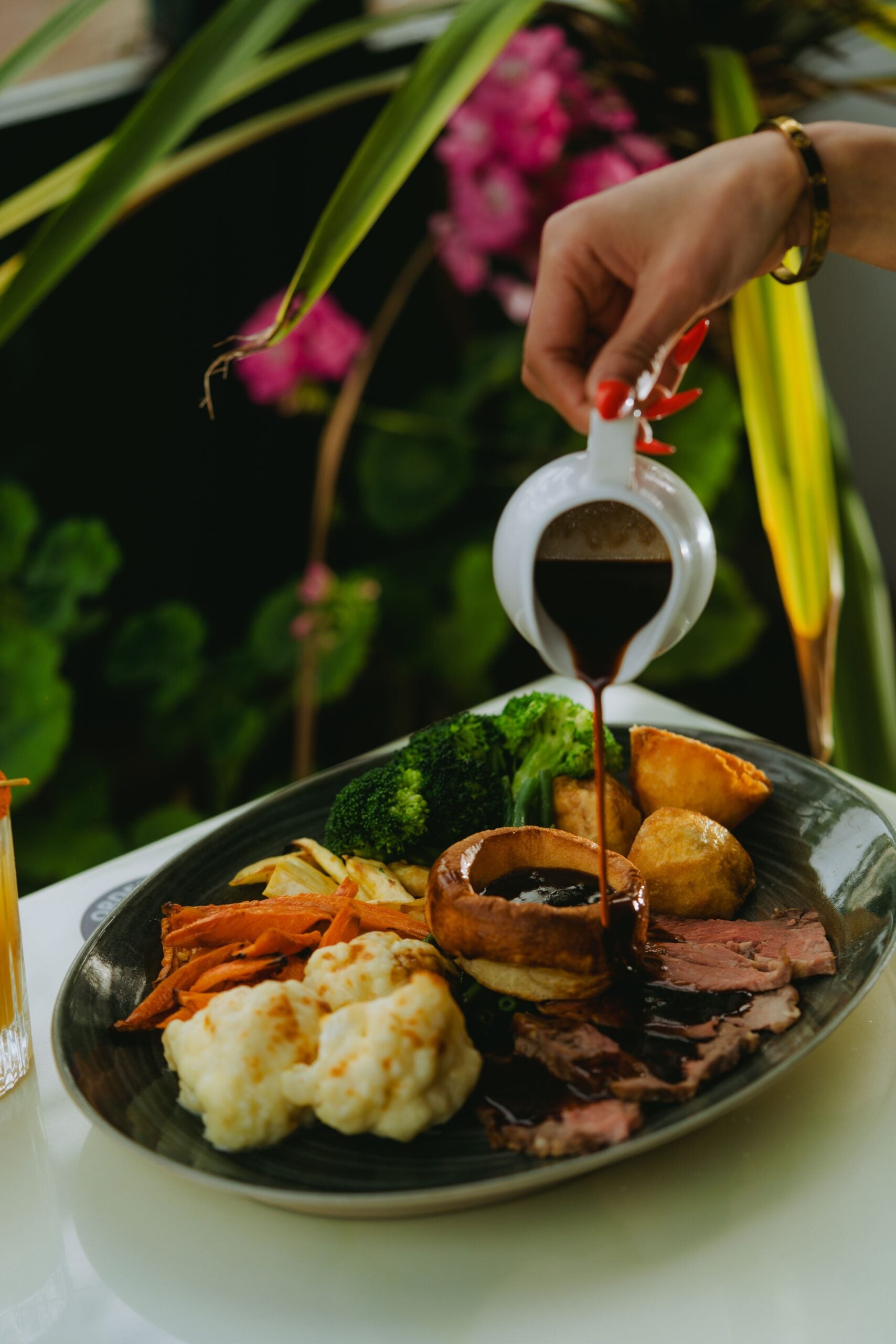 A hand pours brown gravy from a small jug onto a plate of roast beef, Yorkshire pudding, roasted potatoes, broccoli, carrots, and cauliflower. Bright green plants and pink flowers are blurred in the background.