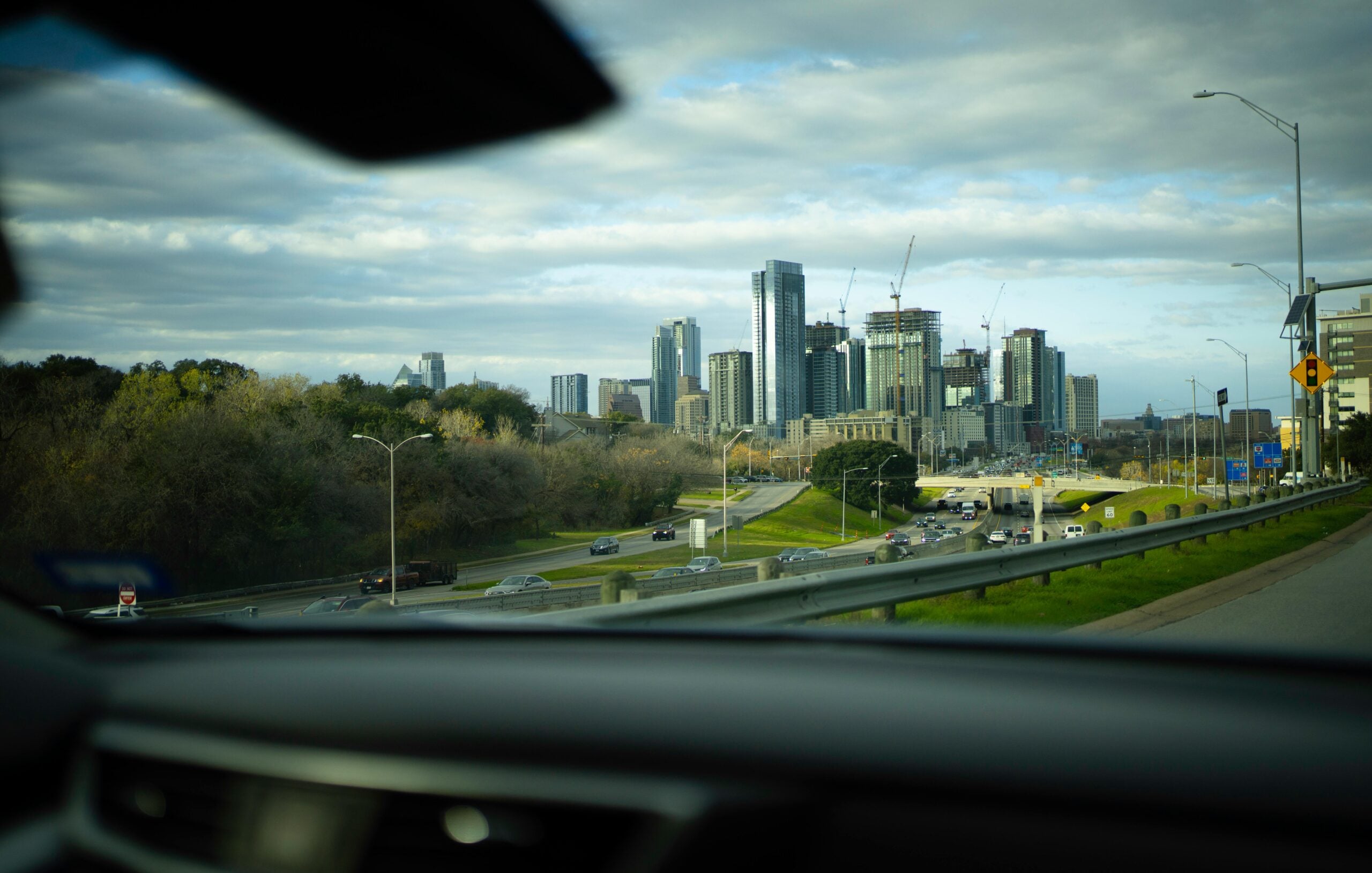 View of a city skyline with tall buildings and cranes in the distance, seen from inside a car driving on a highway with multiple lanes of traffic and trees along the roadside under a partly cloudy sky.