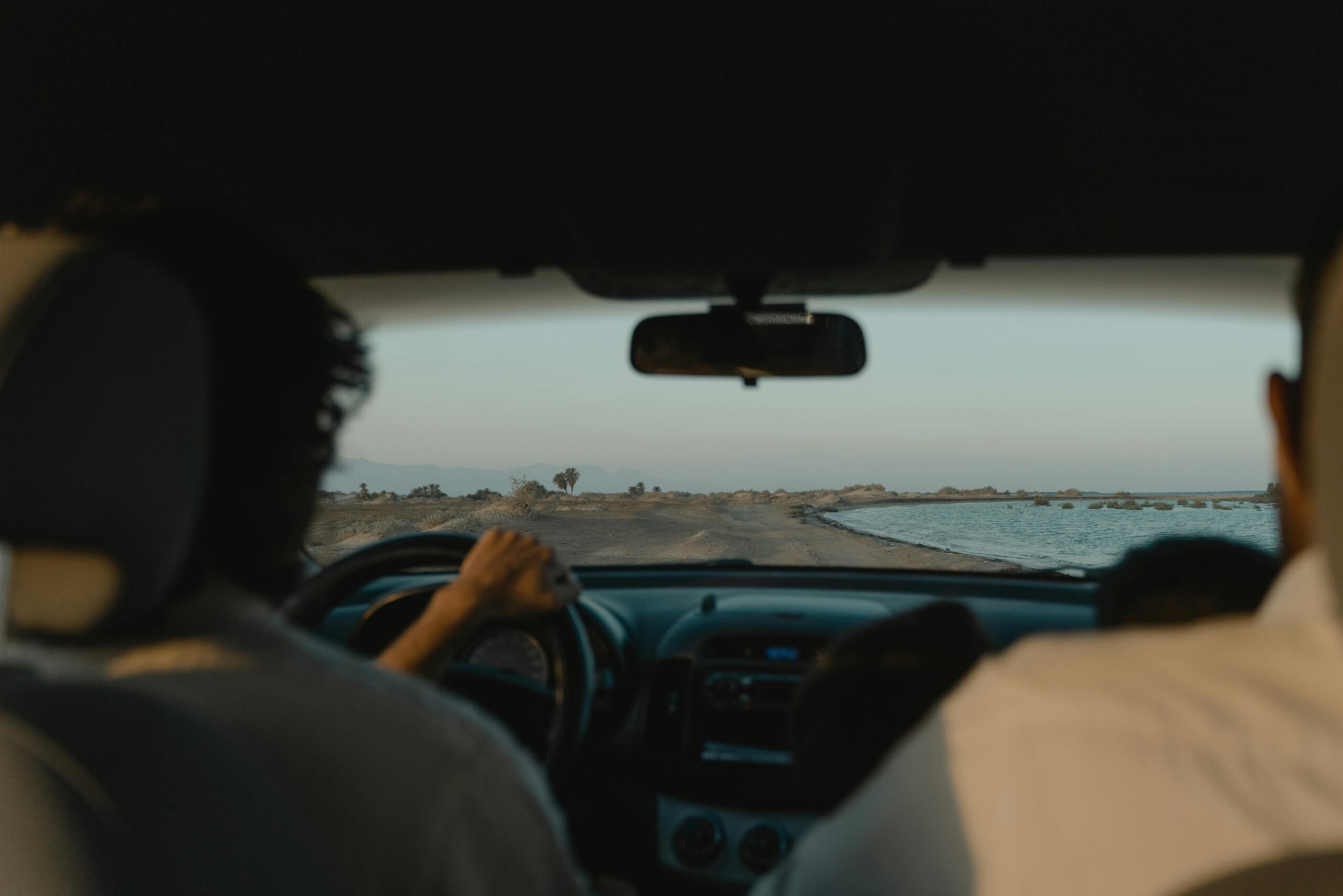 View from inside a car showing two people in the front seats driving along a narrow dirt road beside a body of water, with a clear sky and distant landscape visible through the windshield.