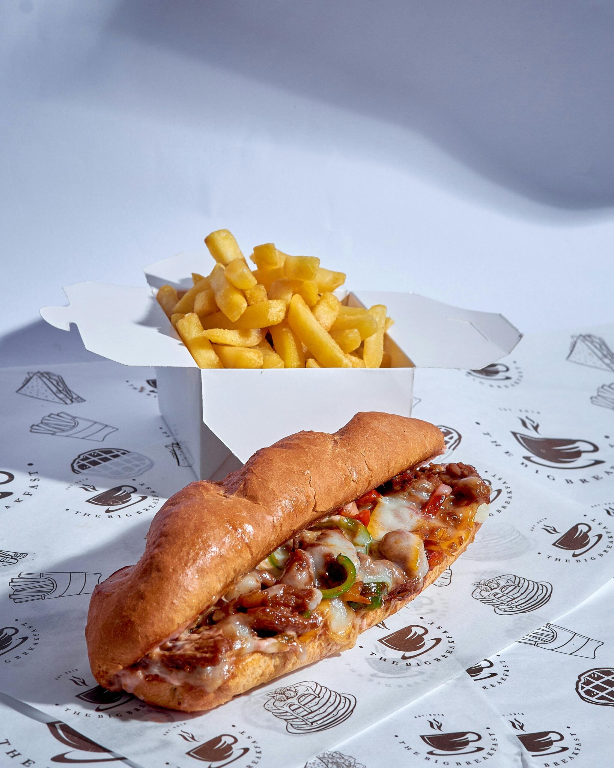 A cheesy sub sandwich filled with vegetables and meat sits on deli paper, with a box of golden French fries in the background.