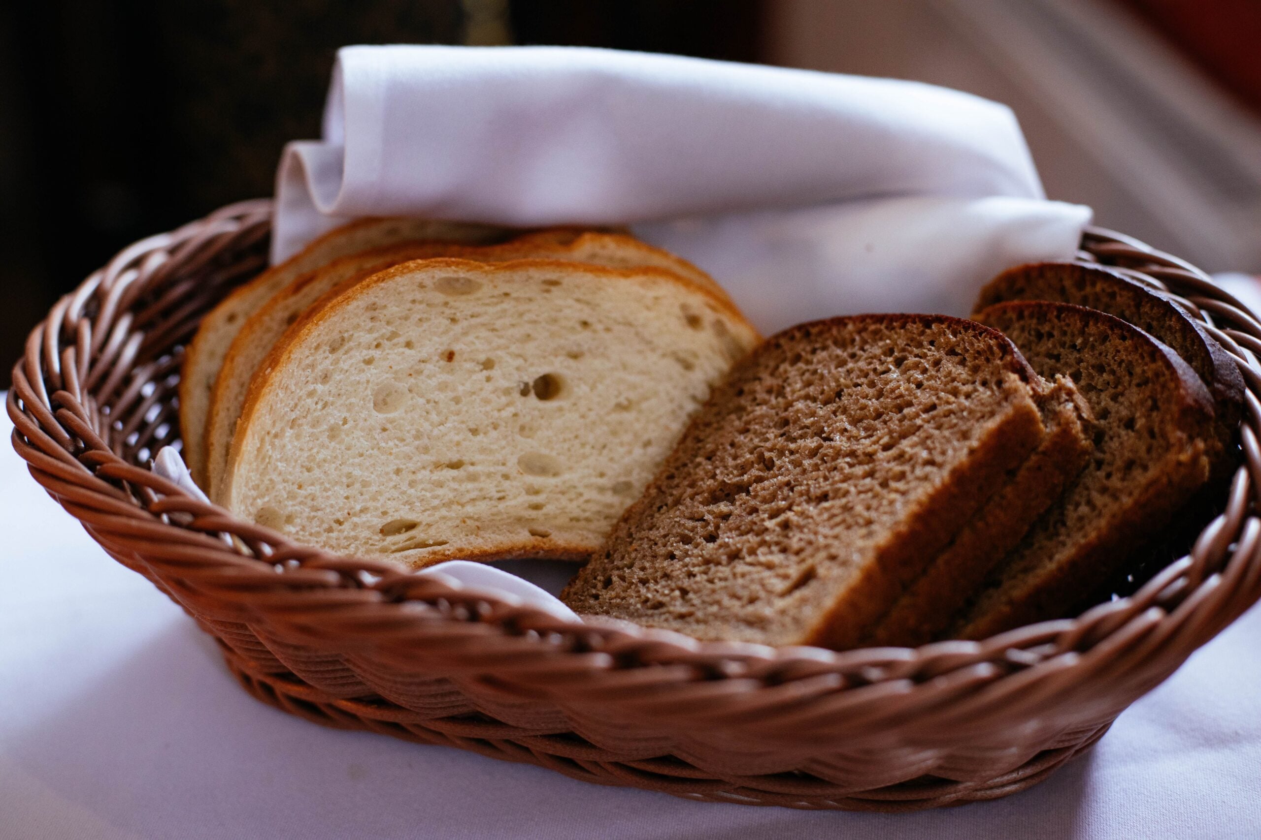 A woven basket lined with a white cloth holds several slices of white bread and brown bread, arranged side by side on a white tablecloth.