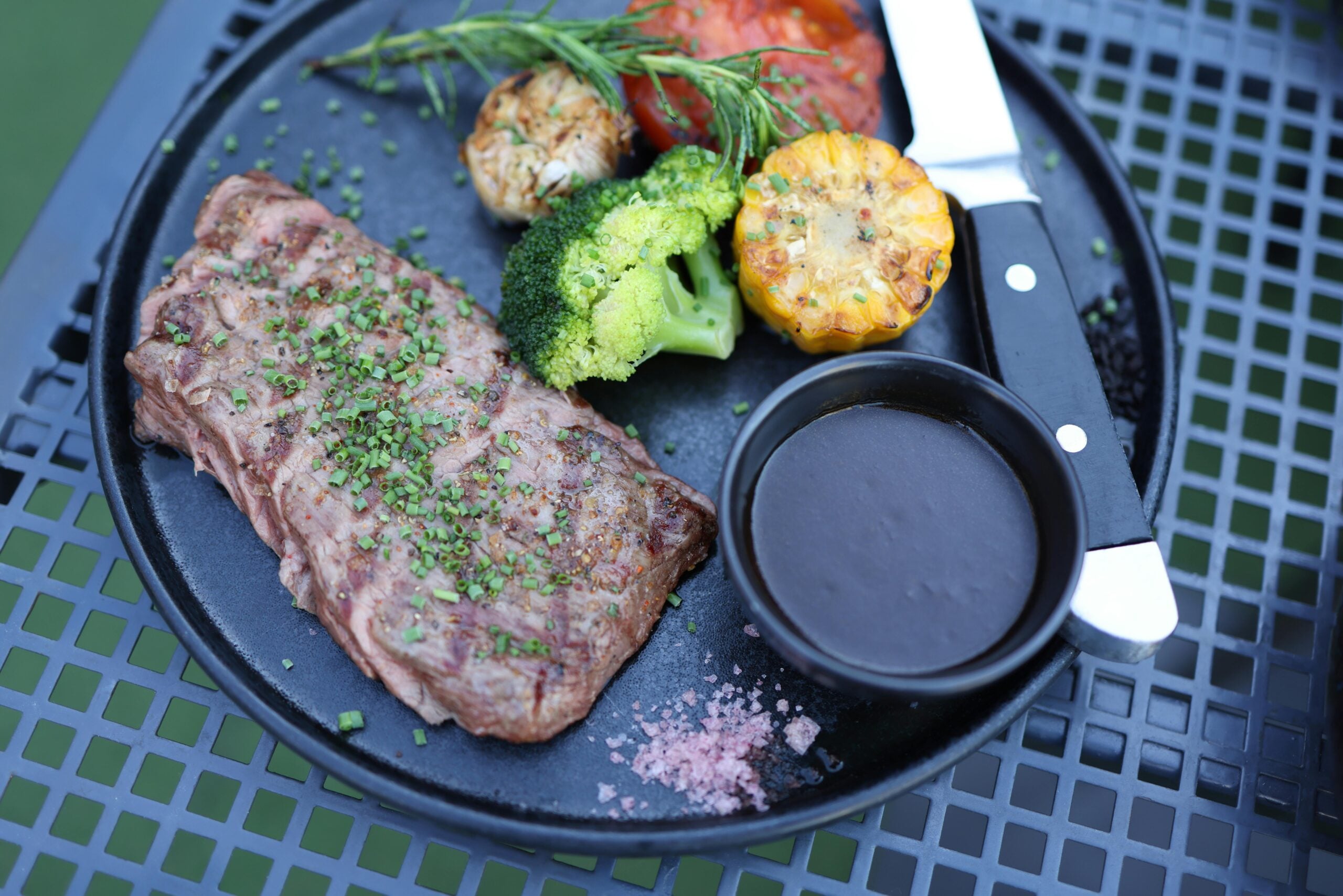 A grilled steak garnished with herbs is served with broccoli, corn, roasted garlic, a tomato, a small dish of dark sauce, and pink salt on a black plate with a knife and fork.