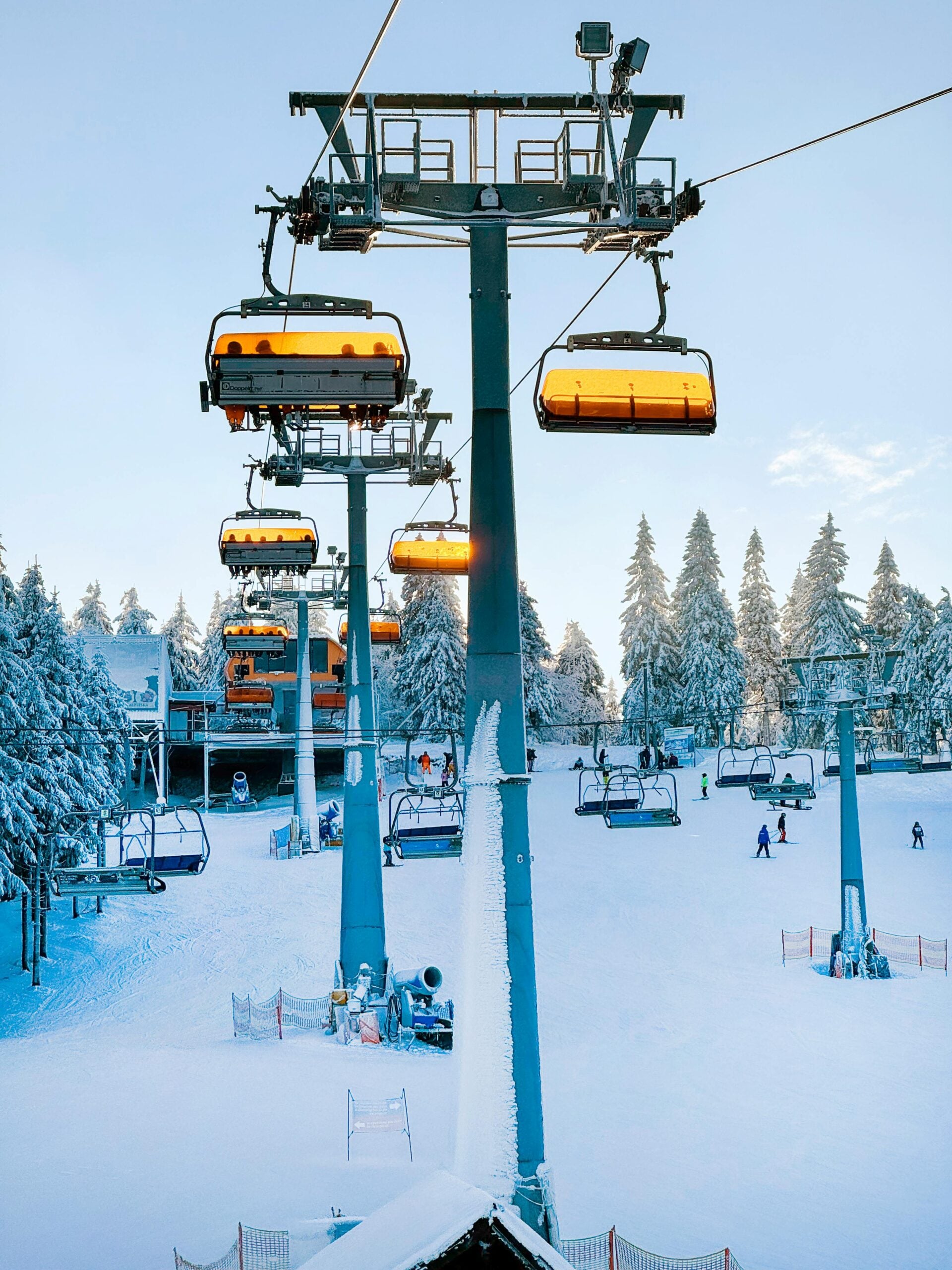 Ski lift chairs with yellow covers move above a snowy ski resort, with skiers and snowboarders on the ground. Snow-covered trees and blue sky are in the background.
