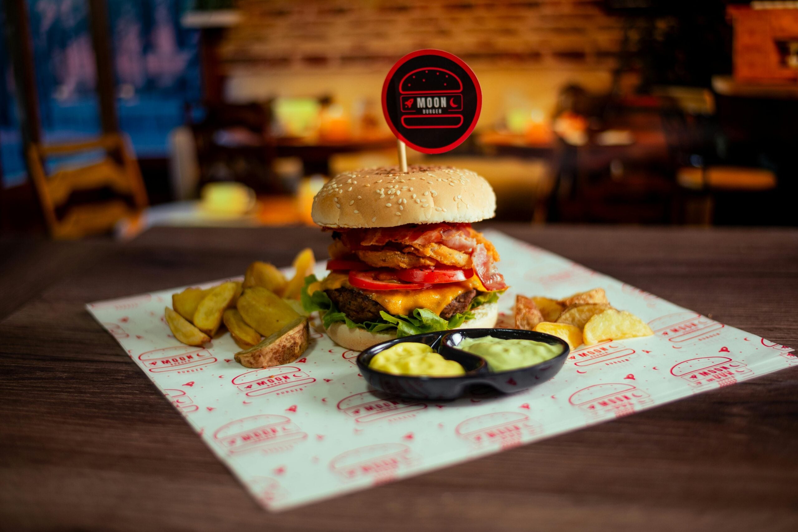 A large burger with bacon, lettuce, tomato, and cheese is served on a tray with thick-cut fries and two dips. The meal is placed on branded paper in a cozy, softly lit restaurant setting.