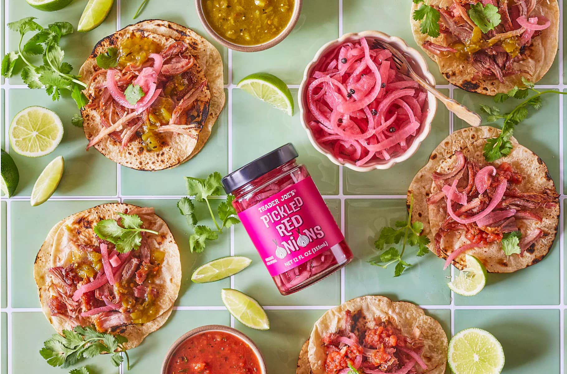 Street tacos topped with pulled meat, pickled red onions, salsa, and cilantro are arranged on green tile with lime wedges, fresh cilantro, salsas, and a jar of pickled red onions in the center.