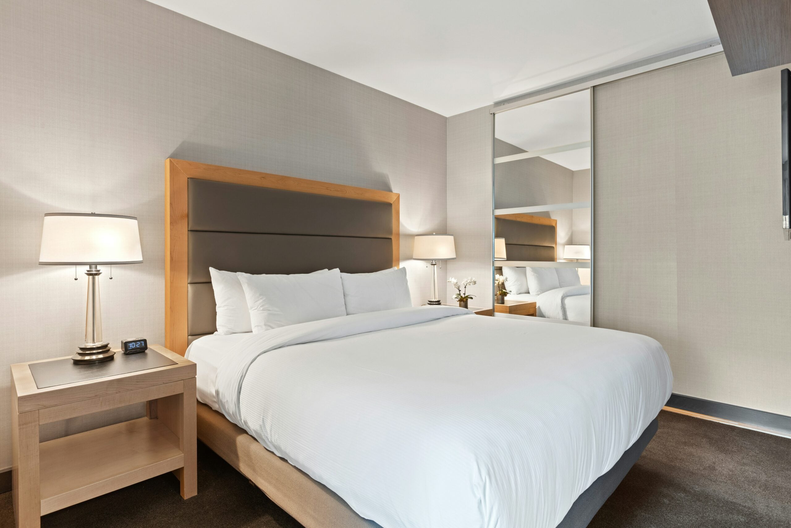 A modern hotel room with a large bed, white bedding, two bedside tables with lamps, a mirrored closet, and neutral-colored walls and carpet.