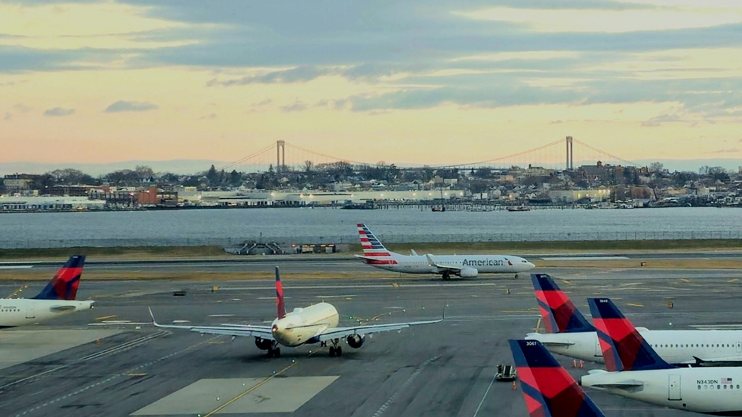 Several airplanes, including Delta and American Airlines, are on a runway at an airport near water. In the background, there is a cityscape and a large suspension bridge under a partly cloudy sky.
