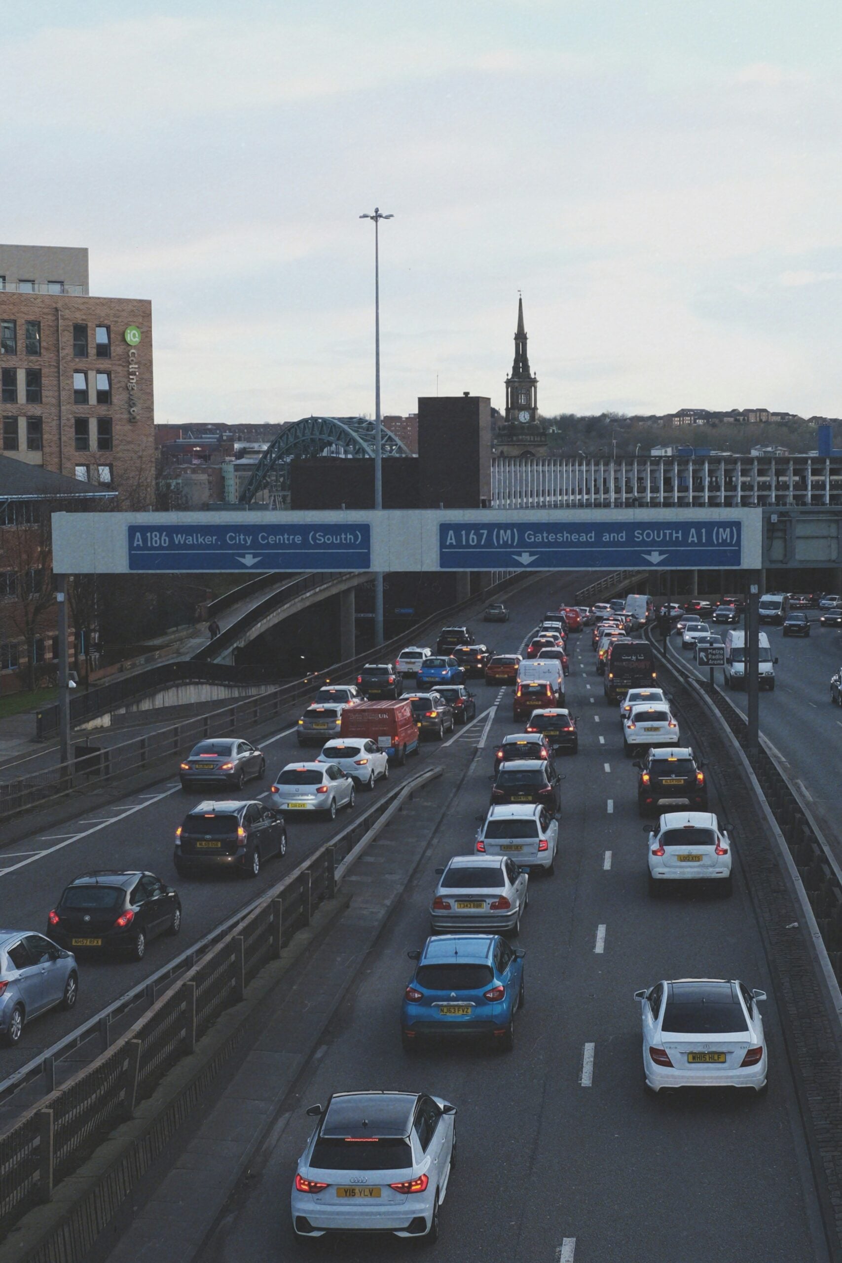 A busy multi-lane motorway with heavy traffic, including cars and vans, under blue directional road signs. City buildings and a church spire are visible in the background under a cloudy sky.