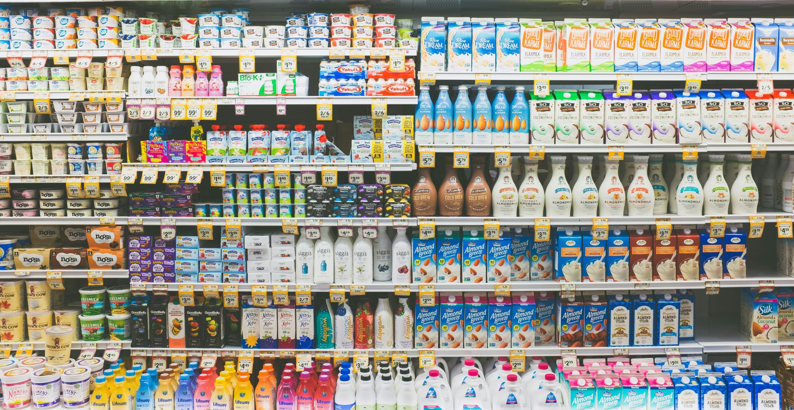 A supermarket refrigerated section stocked with various dairy and non-dairy products, including milk, yogurt, and creamers, organized on shelves with colorful packaging and price tags.