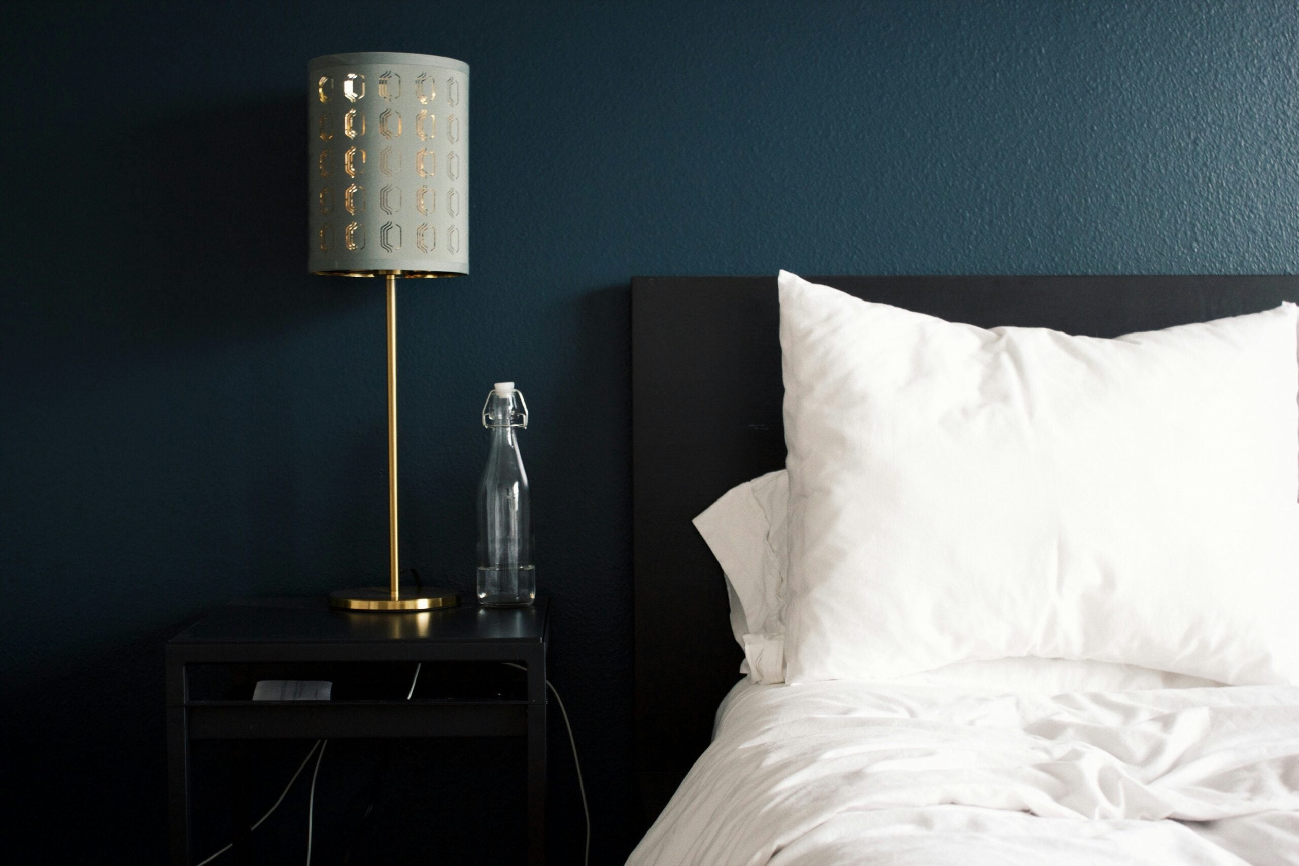 A modern bedroom scene with a dark headboard, white bedding, a black nightstand holding a gold lamp with a patterned shade, and a glass water bottle against a dark blue wall.