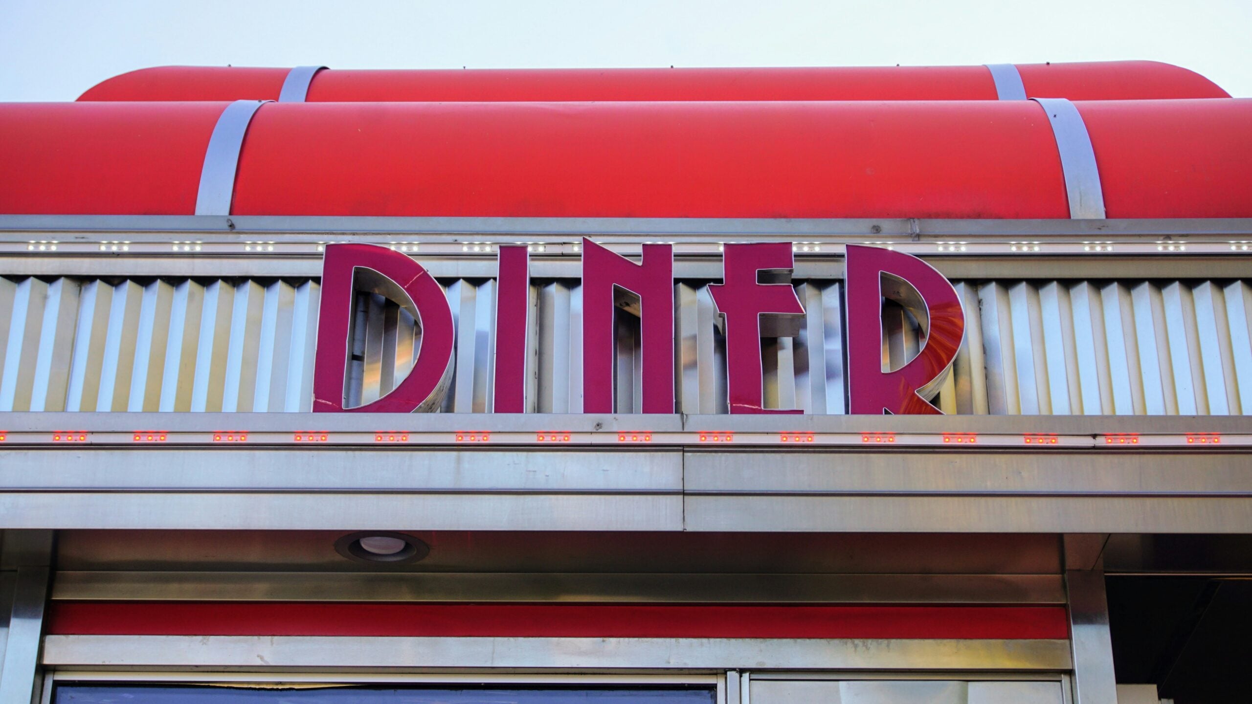 A close-up of a classic diner sign with red letters spelling "DINER" on a silver metallic background and a red curved roof above it against a clear sky.