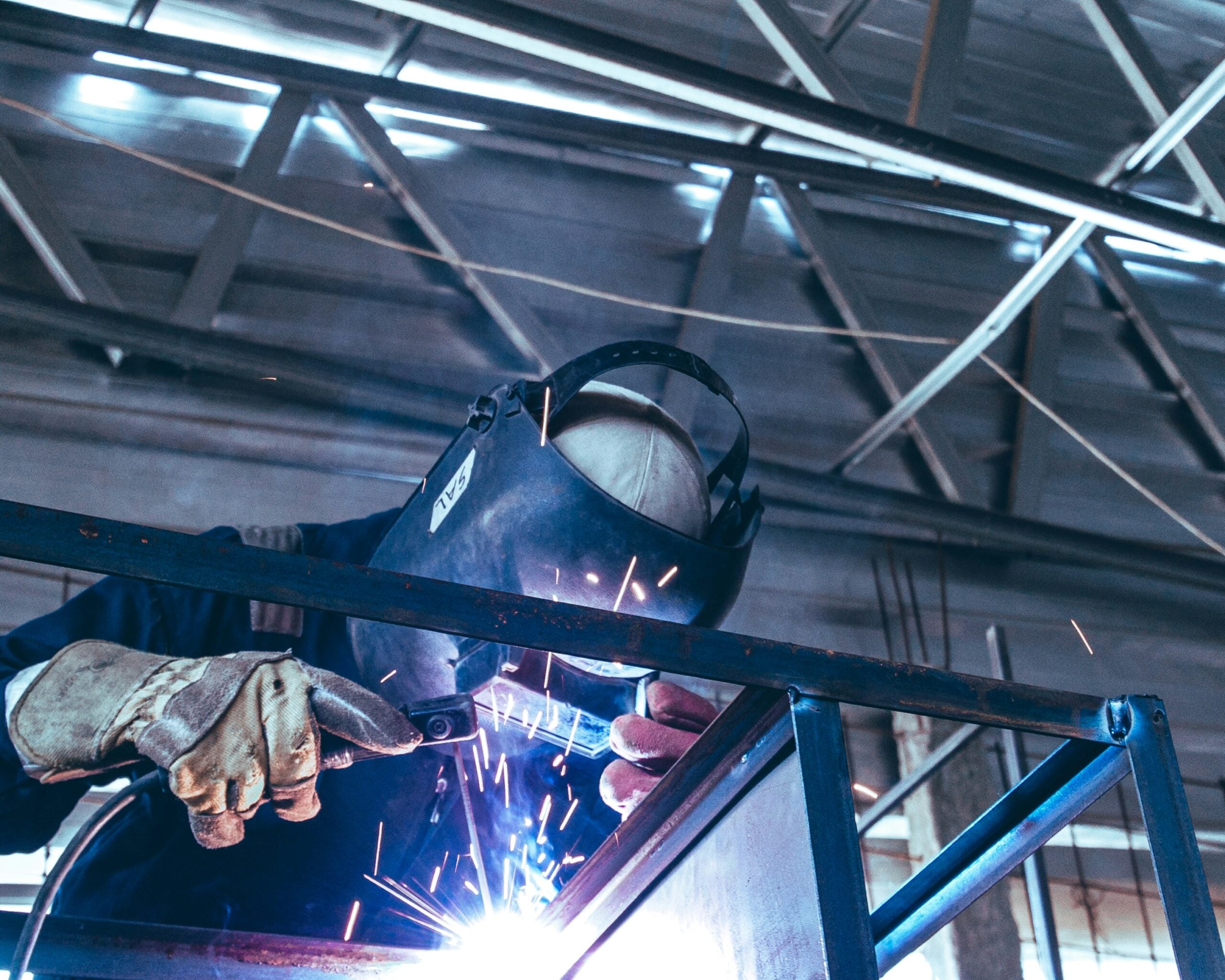 A person wearing protective gloves and a welding helmet is welding metal beams indoors, with bright sparks flying from the welding torch. Structural metal beams and a high ceiling are visible in the background.