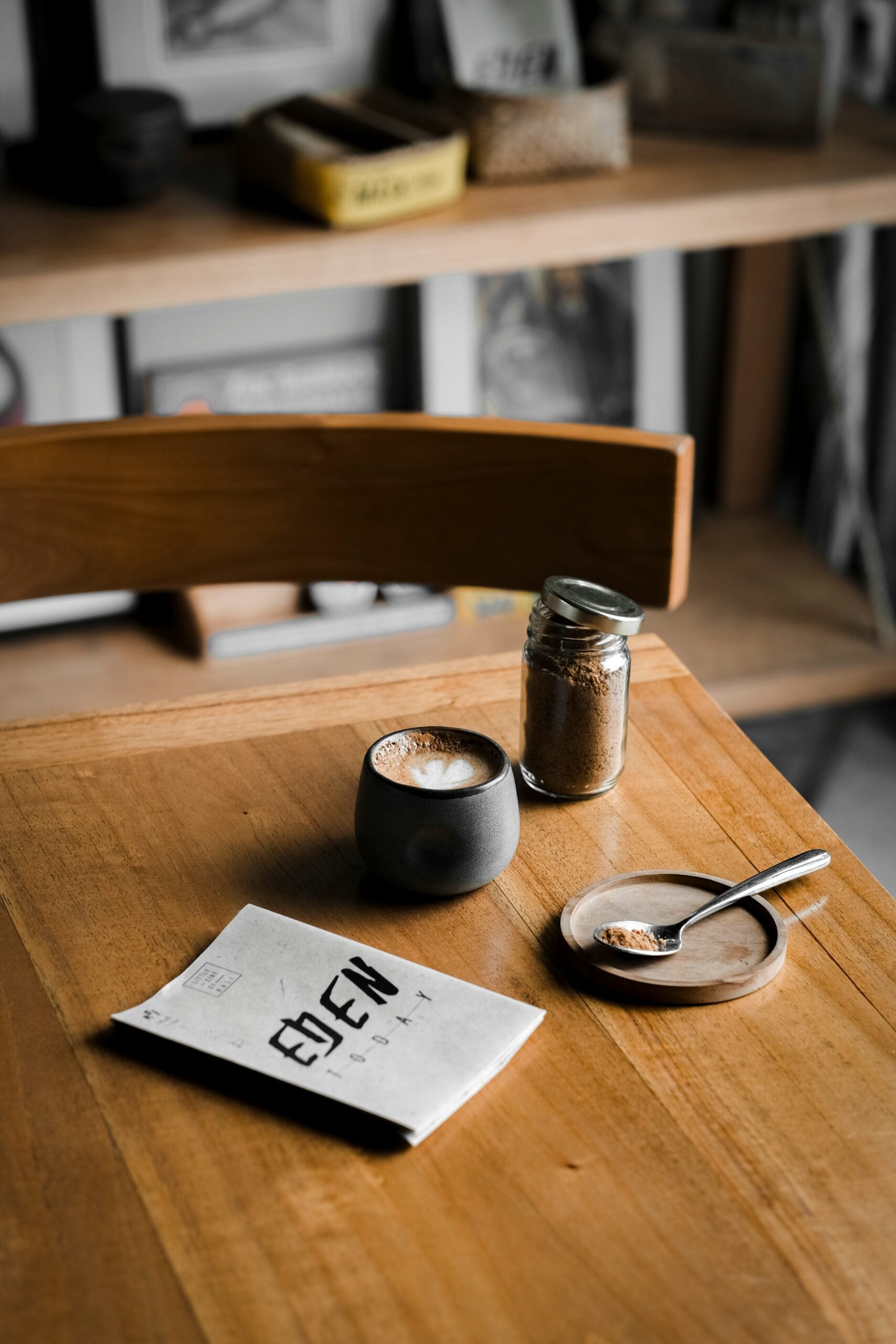 A wooden table with a cup of coffee, a small jar of sugar, a spoon on a dish, and a folded menu labeled “EDEN.” Shelves with books and containers are blurred in the background.