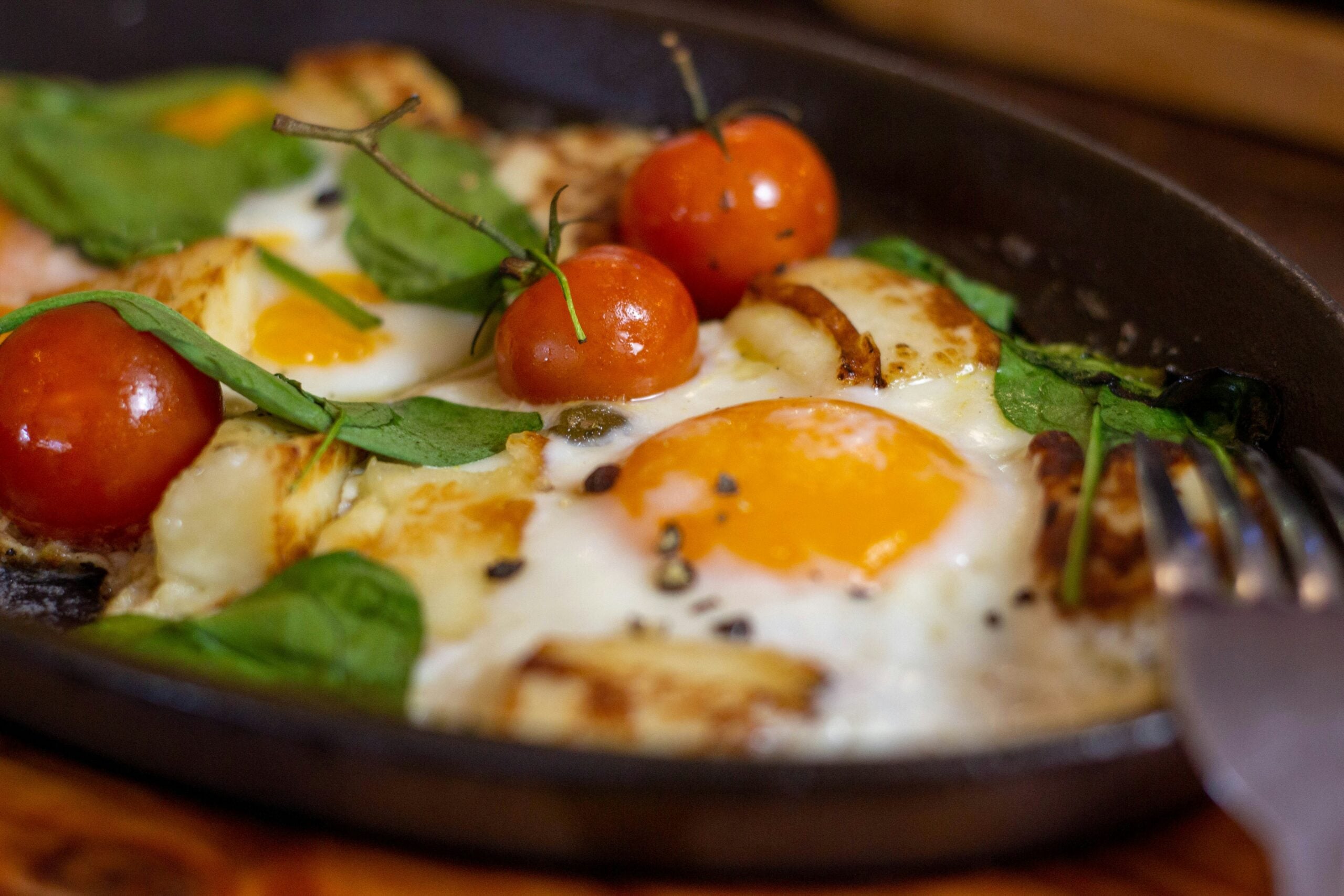 A close-up of a skillet dish with sunny-side-up eggs, cherry tomatoes on the vine, wilted spinach, and melted cheese, garnished with black pepper. A fork rests at the edge of the pan.