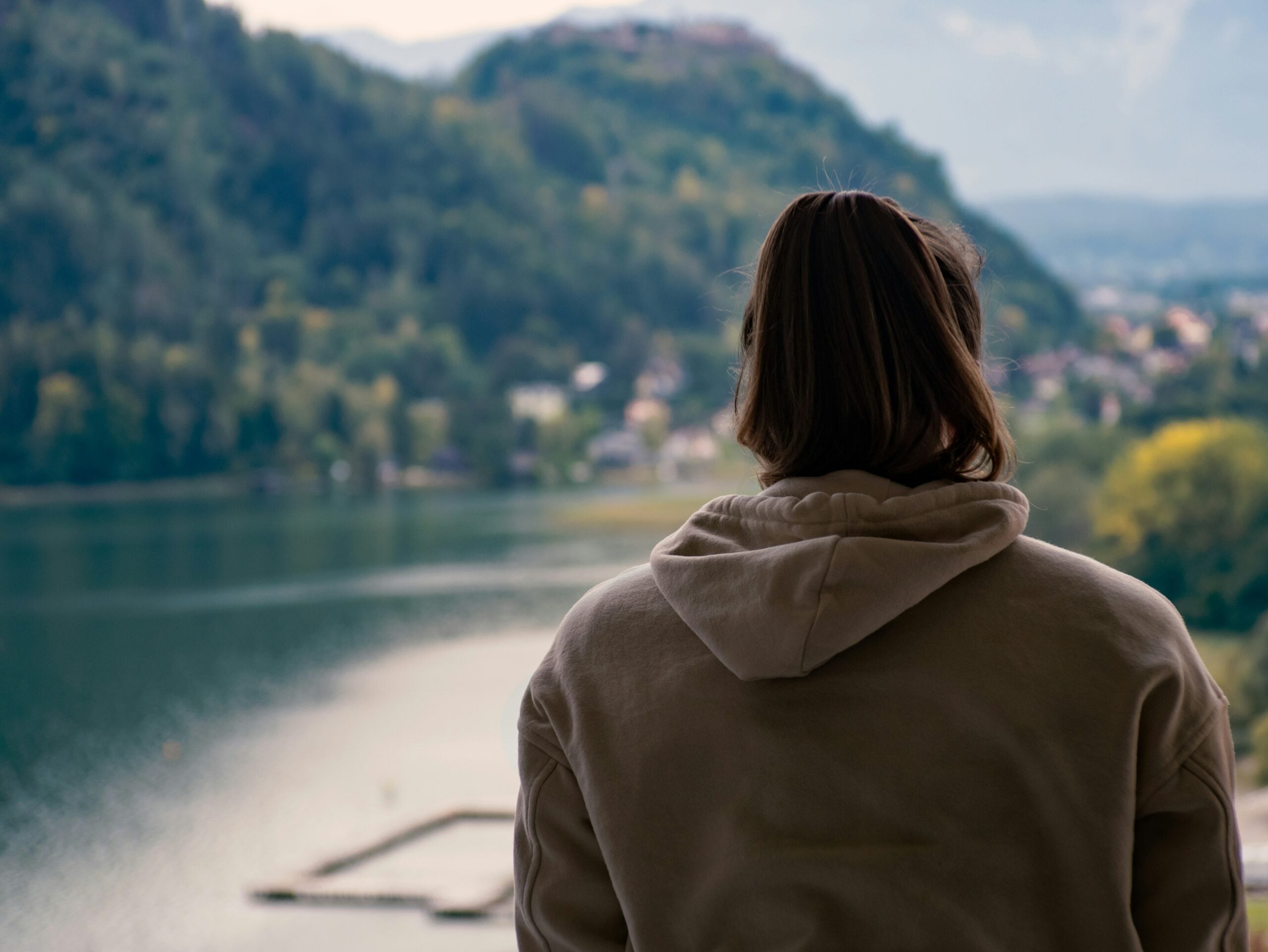A person with shoulder-length hair wearing a light hoodie stands with their back to the camera, looking out over a lake surrounded by green hills and trees.
