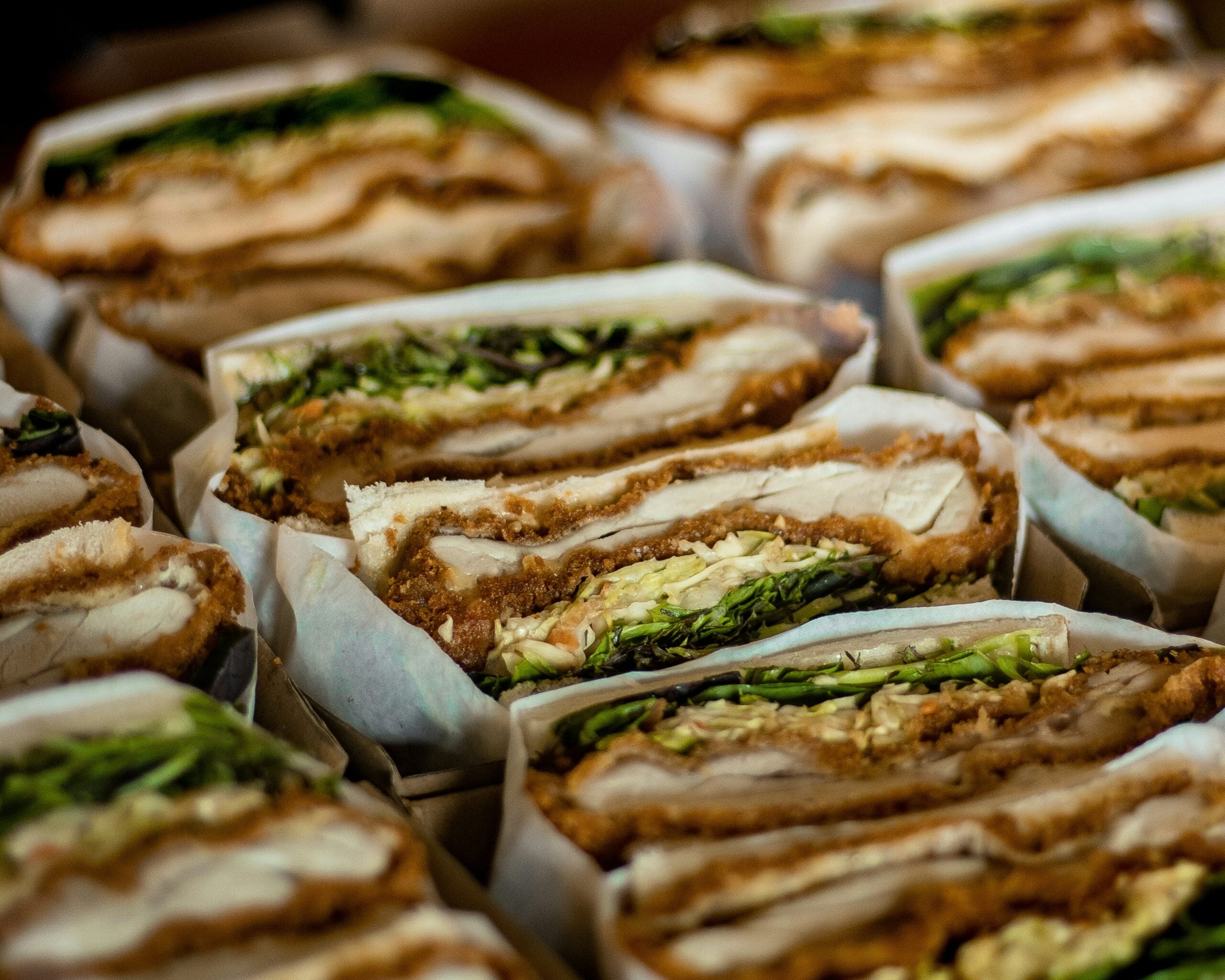 Close-up of several stacked sandwiches, each filled with layers of crispy fried chicken, lettuce, and thinly sliced vegetables, wrapped partially in white paper.