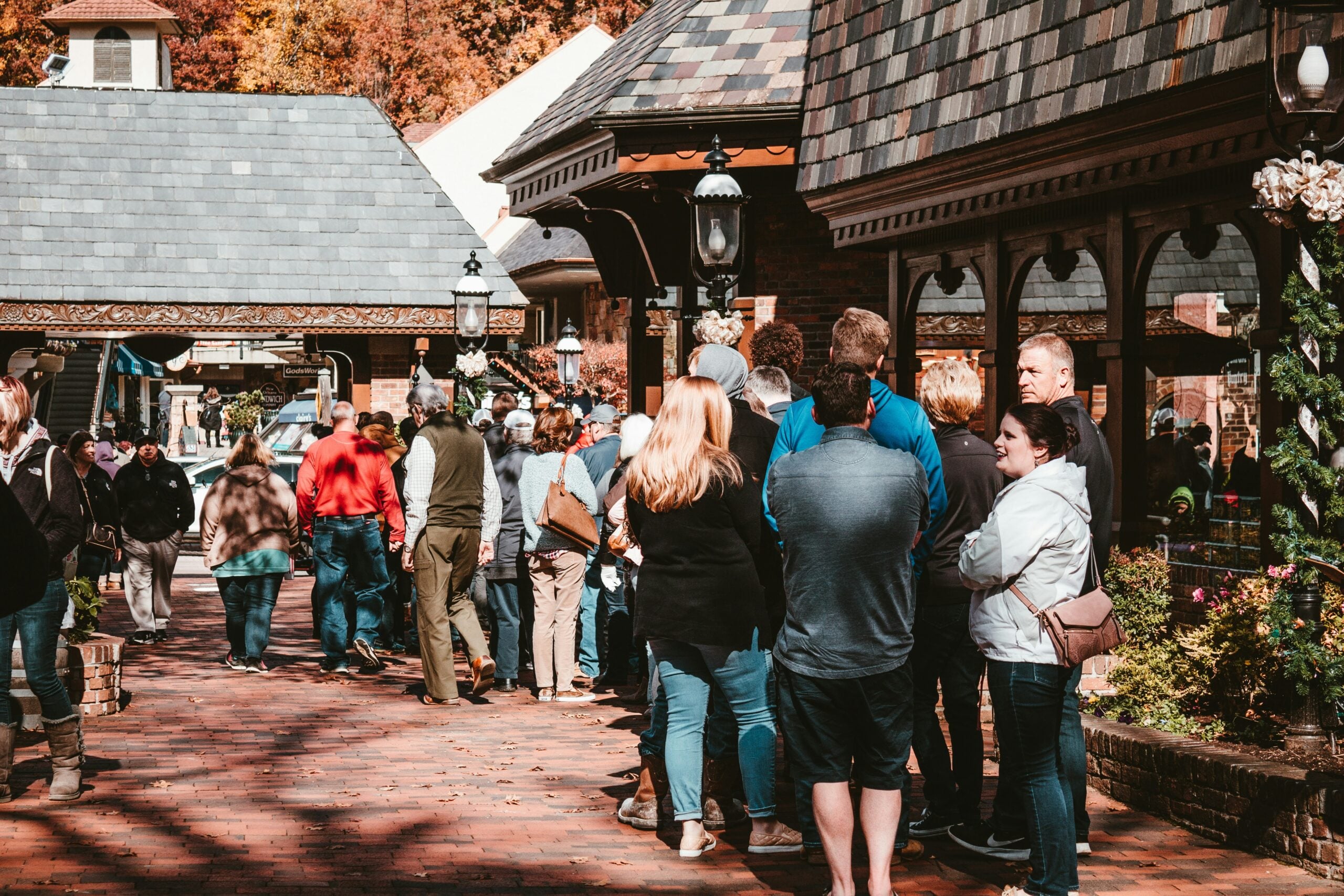 A long line of people stands outside on a brick walkway between shops, waiting in cool weather. Some are dressed in jackets and sweaters. Trees with autumn leaves are visible in the background.