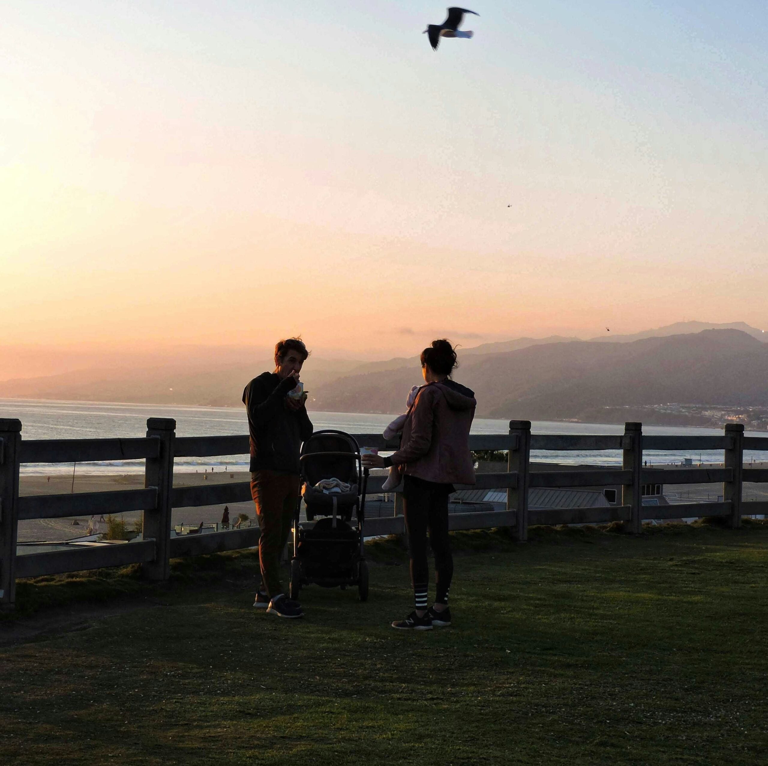 Two people stand beside a stroller on a grassy area near a wooden fence, overlooking the ocean at sunset. Mountains are visible in the background, and a bird flies overhead in the sky.