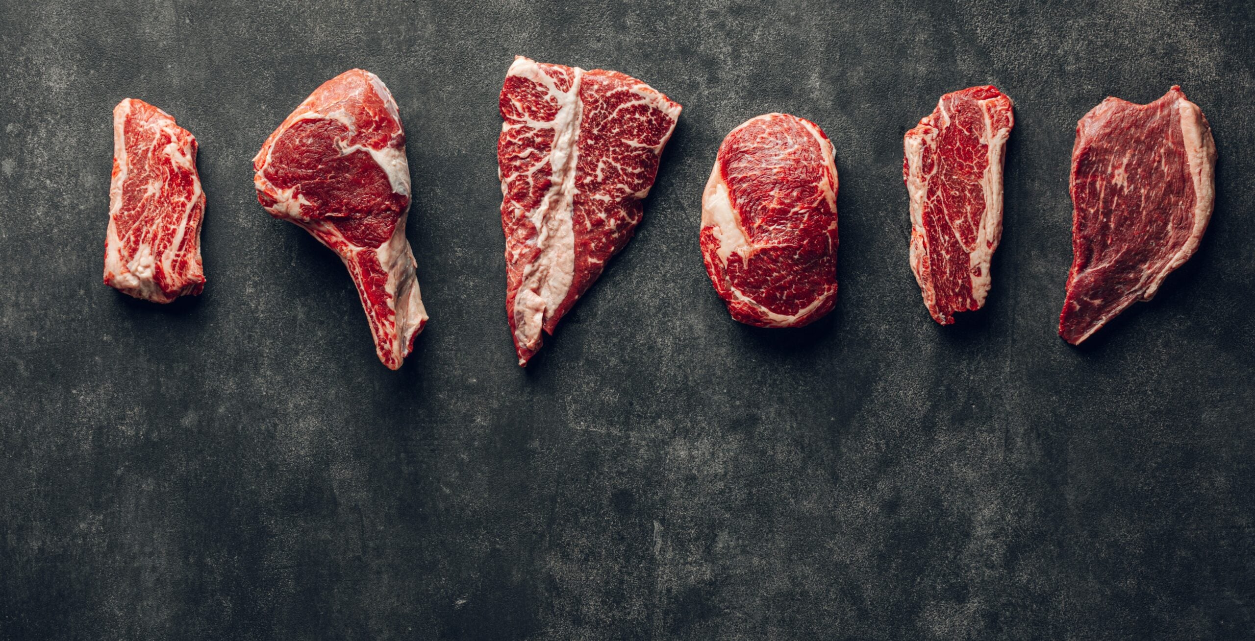 Six different raw beef steaks are arranged in a row on a dark, textured surface, showcasing various cuts with visible marbling and fat.