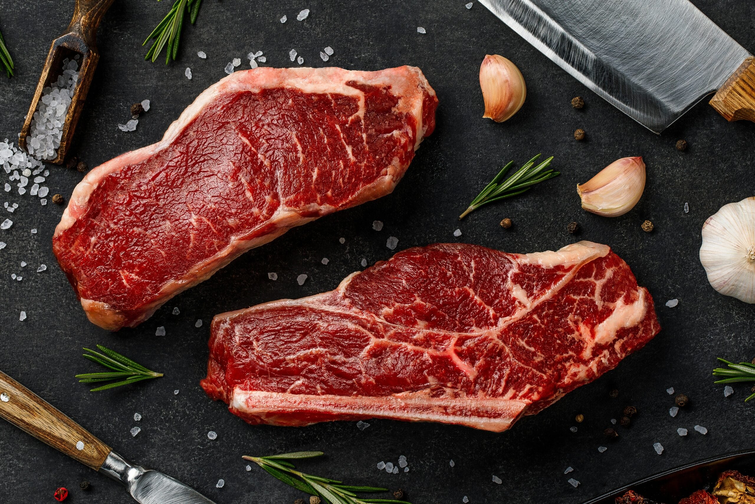 Two raw steaks on a dark surface, surrounded by sprigs of rosemary, garlic cloves, black peppercorns, coarse salt, a meat cleaver, and a fork. The ingredients are arranged for seasoning and preparing the meat.