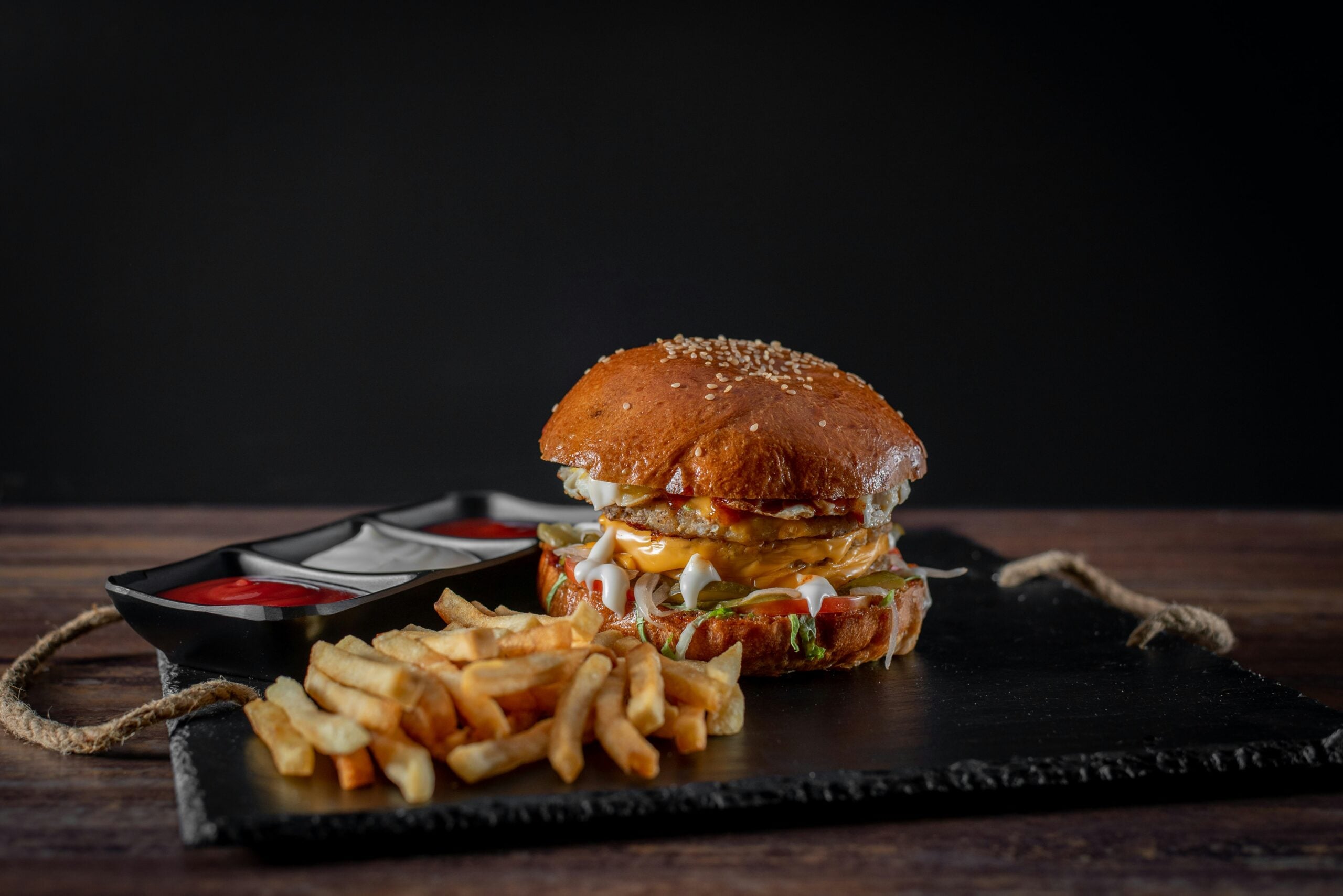 A cheeseburger with lettuce, tomato, cheese, and sauce is served on a black slate with a side of golden French fries and two small containers of ketchup and mayonnaise, all set against a dark background.