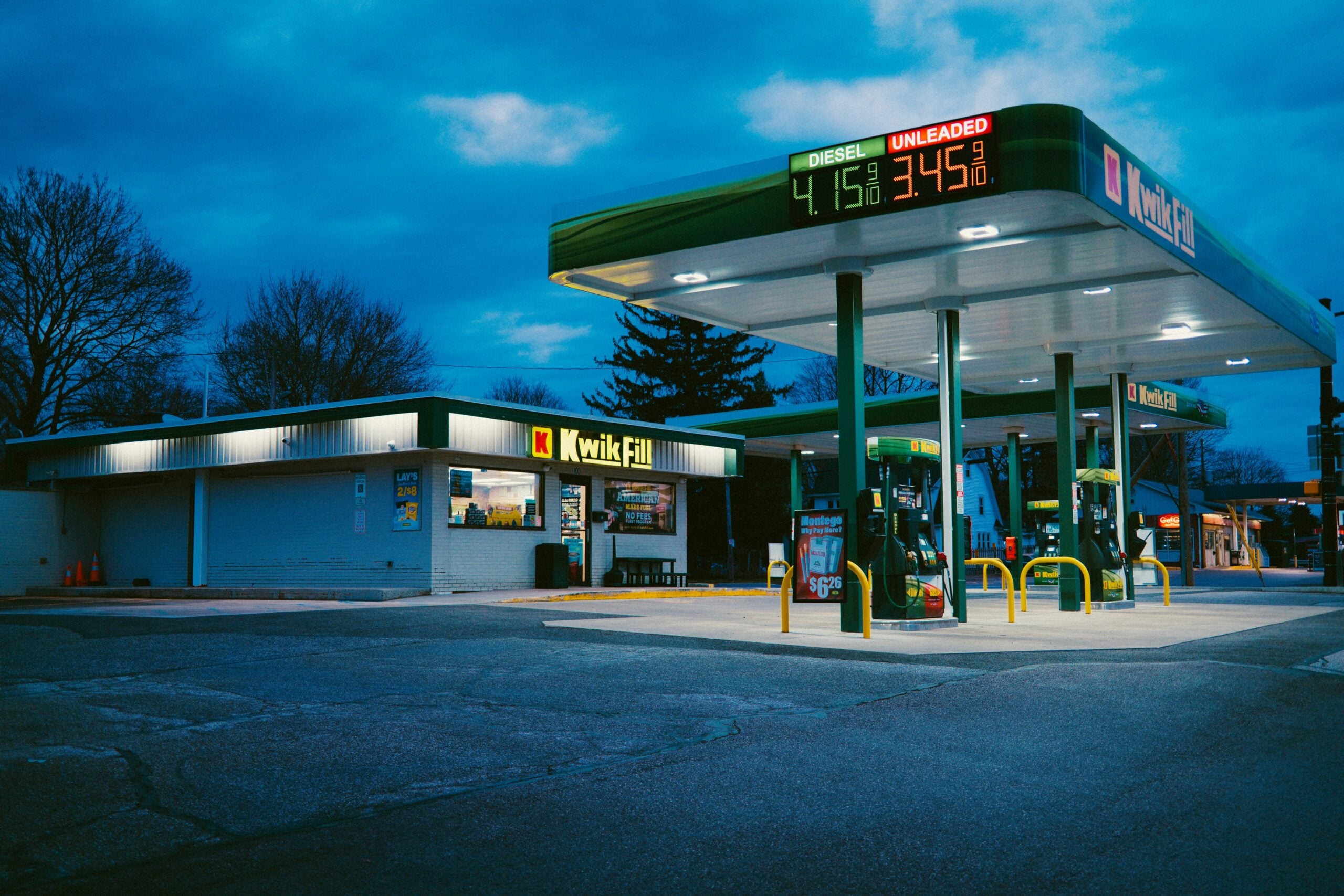 A Kwik Fill gas station at dusk with illuminated signs showing diesel at $4.15 and unleaded at $3.45. The station is mostly empty, with a convenience store in the background and a darkening blue sky.