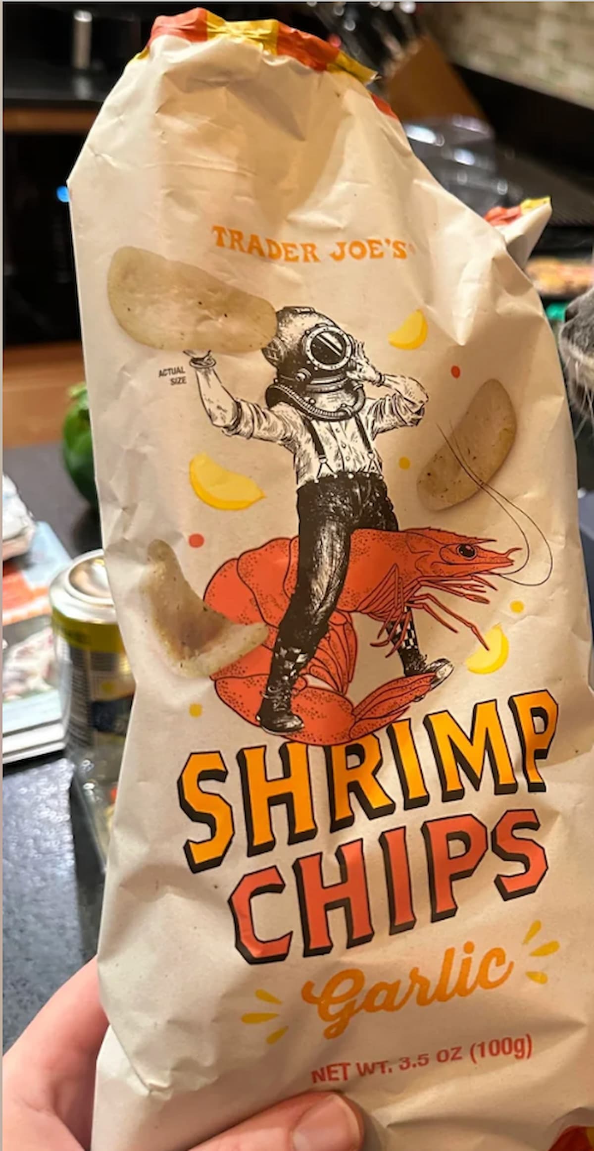 A bag of Trader Joe’s Shrimp Chips, Garlic flavor, featuring an illustration of a person in a vintage diving suit standing on a shrimp, with garlic cloves and chips around them. Net weight 3.5 oz (100g).