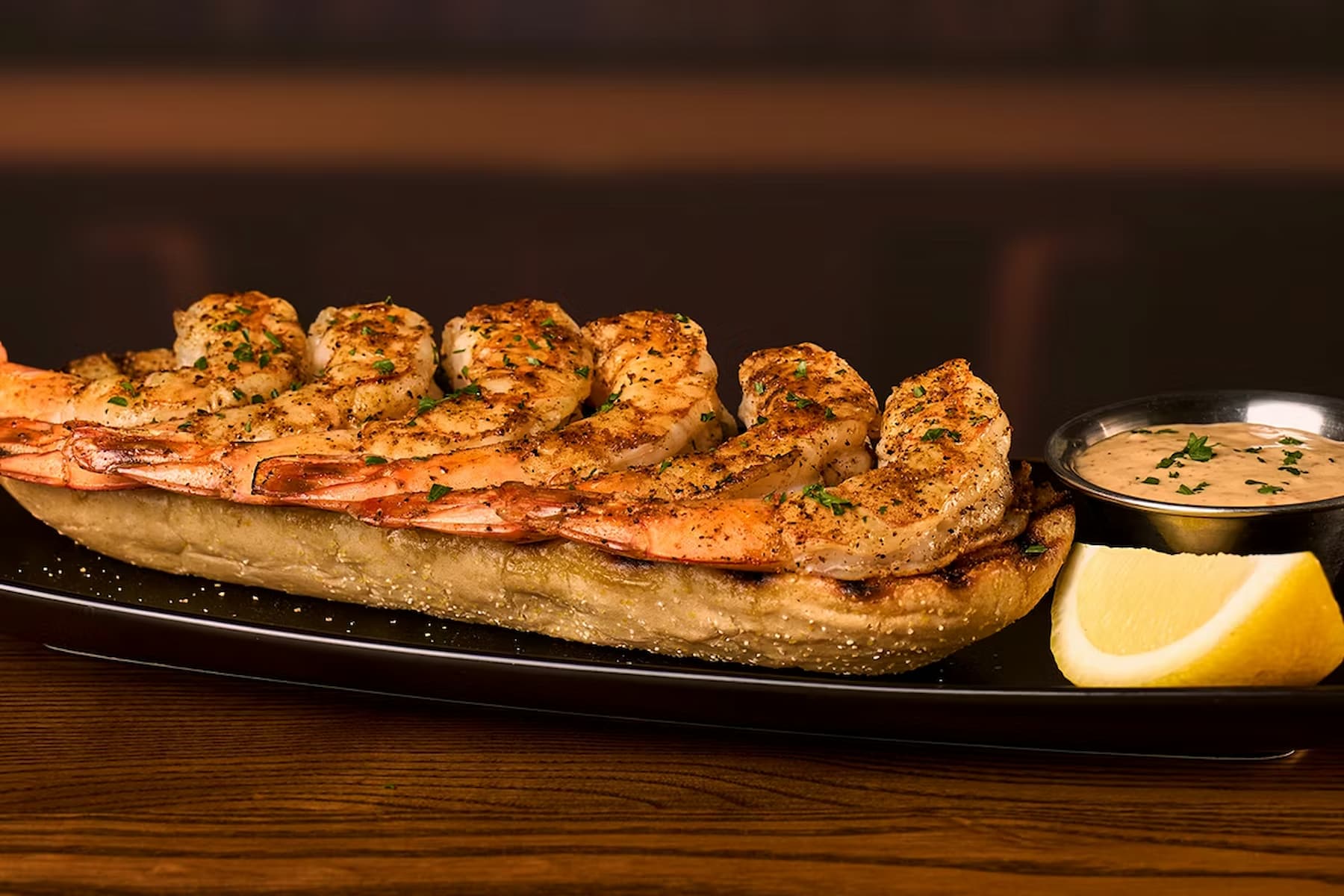 Grilled shrimp skewers served on a toasted baguette with a side of creamy dipping sauce and a lemon wedge, presented on a black rectangular plate.