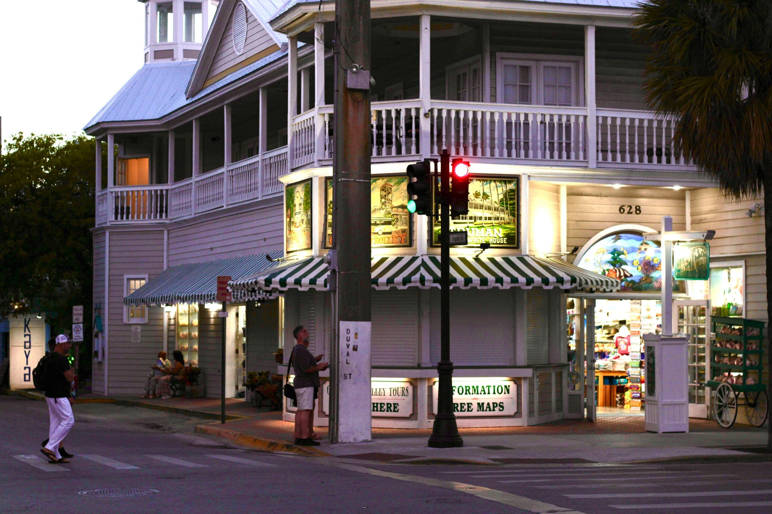 Two people stand on a street corner near a tourist information shop with green-and-white striped awnings, bright lights inside, and a balcony above. A traffic light glows red in front of the wooden building.